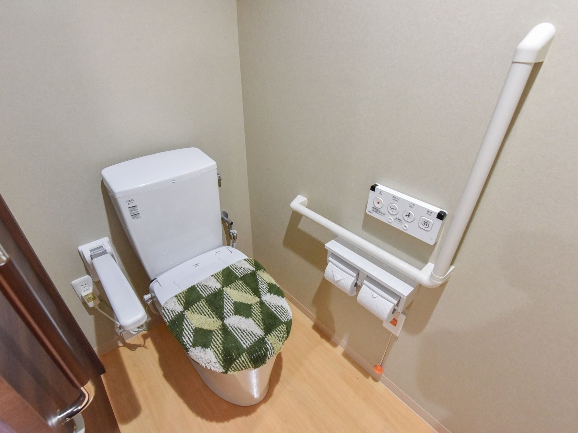 温水洗浄便座が設置され、ふたにはカバーが掛けられている。横には折り畳み式の手すりがついており、壁にはL字型の手すりや操作ボタンなどがある。