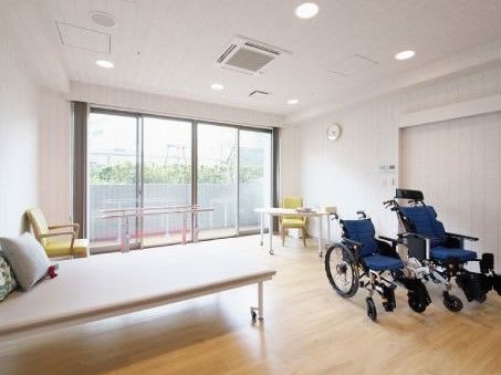 フローリングの床面に、白で統一された壁面が組み合わさった明るい空間である。室内には、歩行用の器具やマッサージ台などの設備が用意されている。