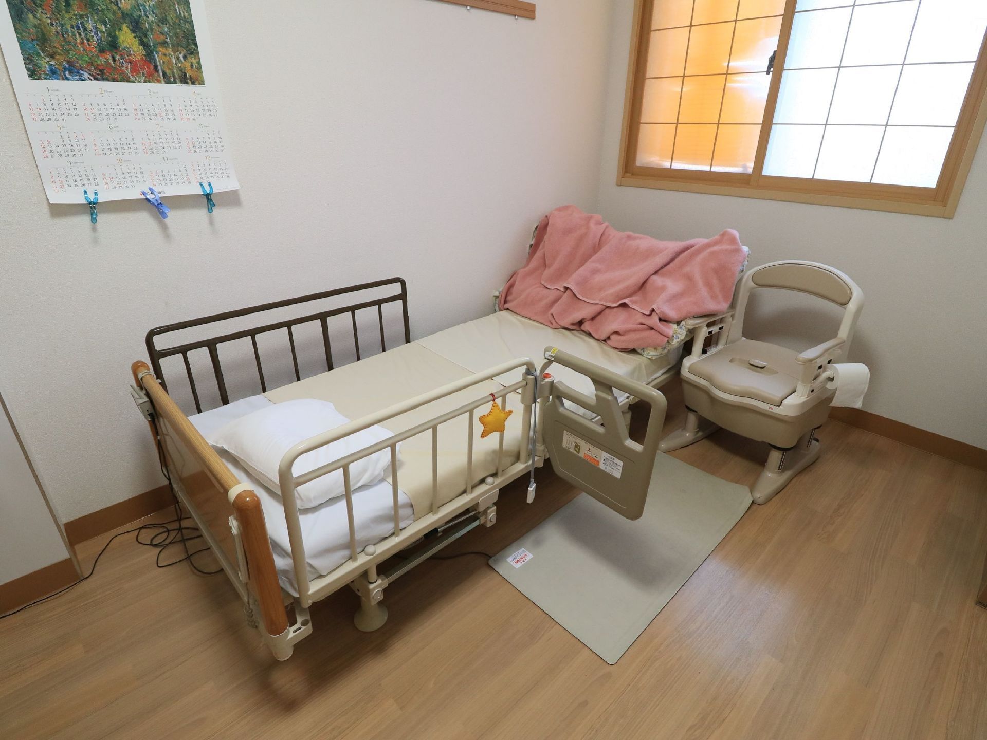 リモコン式の電動介護ベッドの横に、簡易トイレが置かれている。ベッドから降りる際に滑らないよう、滑り止めマットが敷かれている。