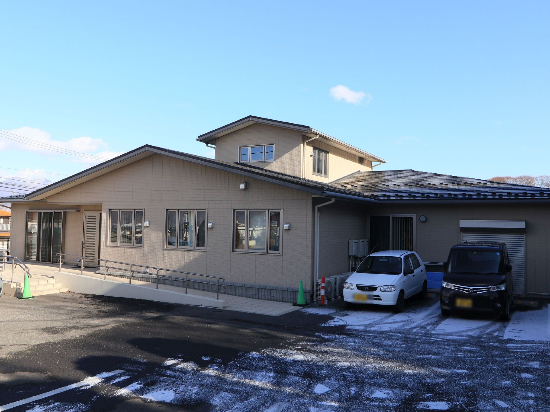 ベージュ色の外壁に三角屋根の二階建ての建物である。玄関前にスロープがあり、駐車スペースも備わっている。
