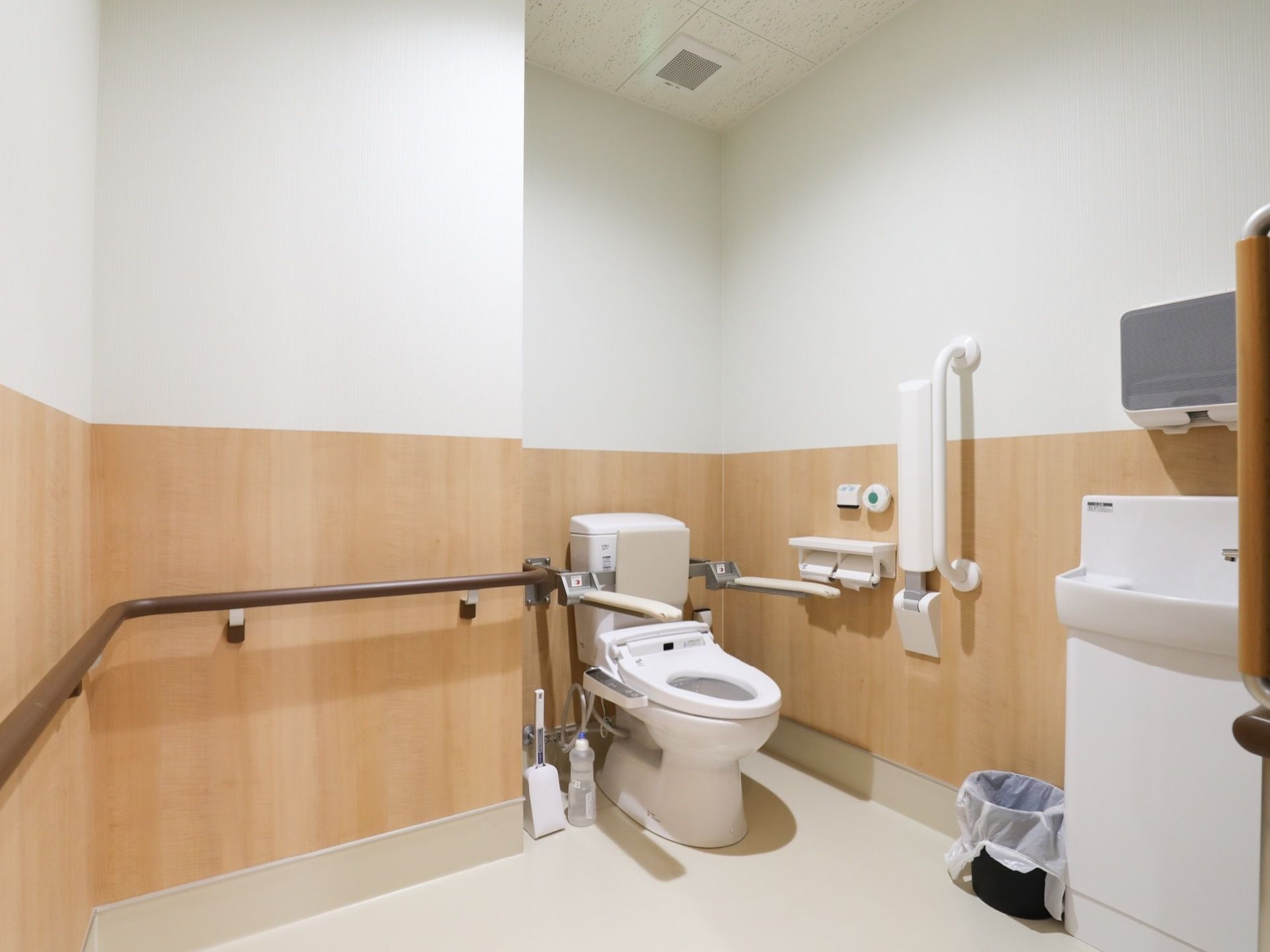 温水洗浄機能があるひじ掛け付きのトイレで、壁には手すりが取り付けてある。手洗い場が設置された広々とした個室である。
