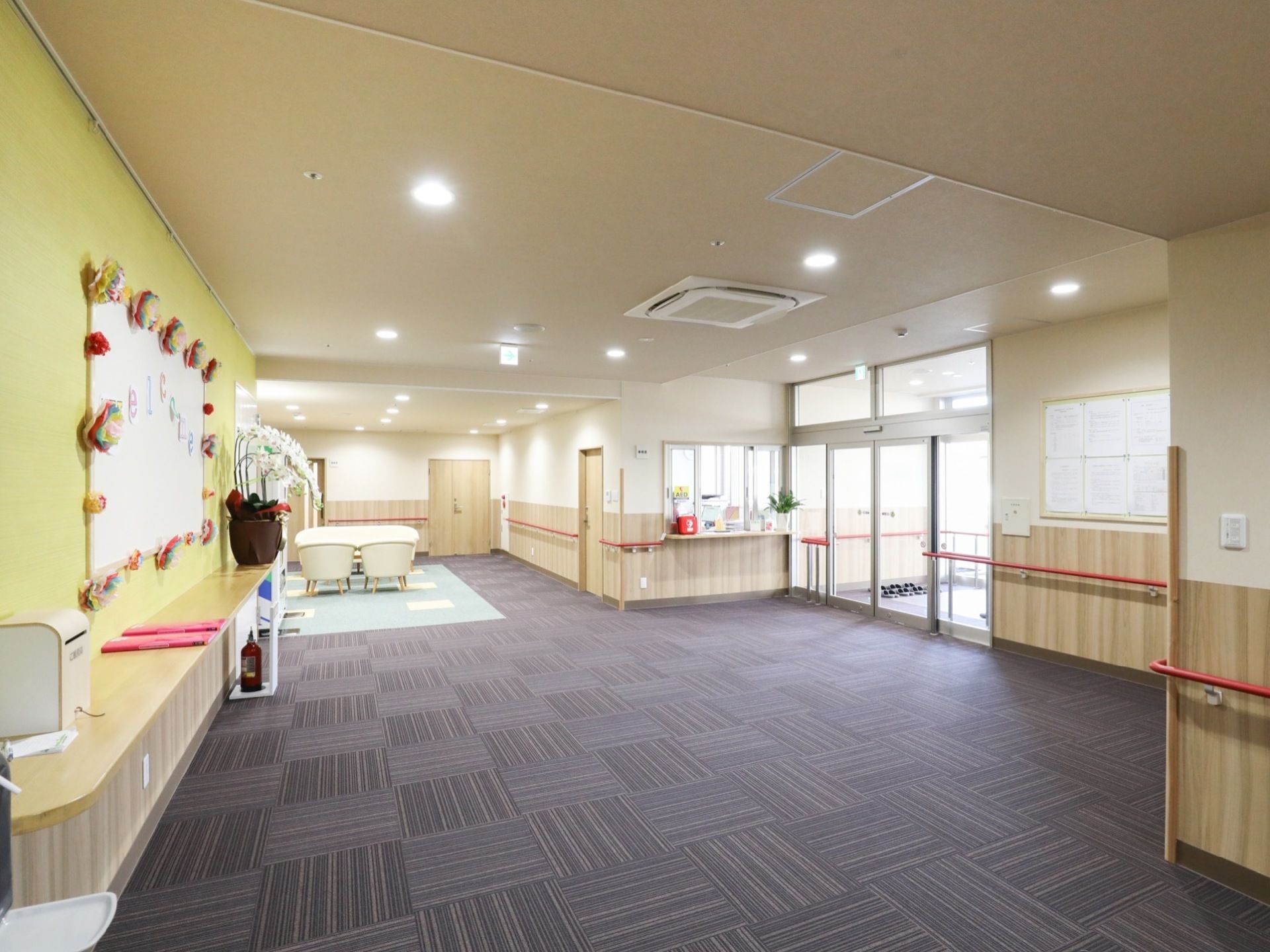 自動ドアの横にガラス窓のついた事務室がある。広々とした玄関ホールで、壁には掲示板やホワイトボードが設置されている。