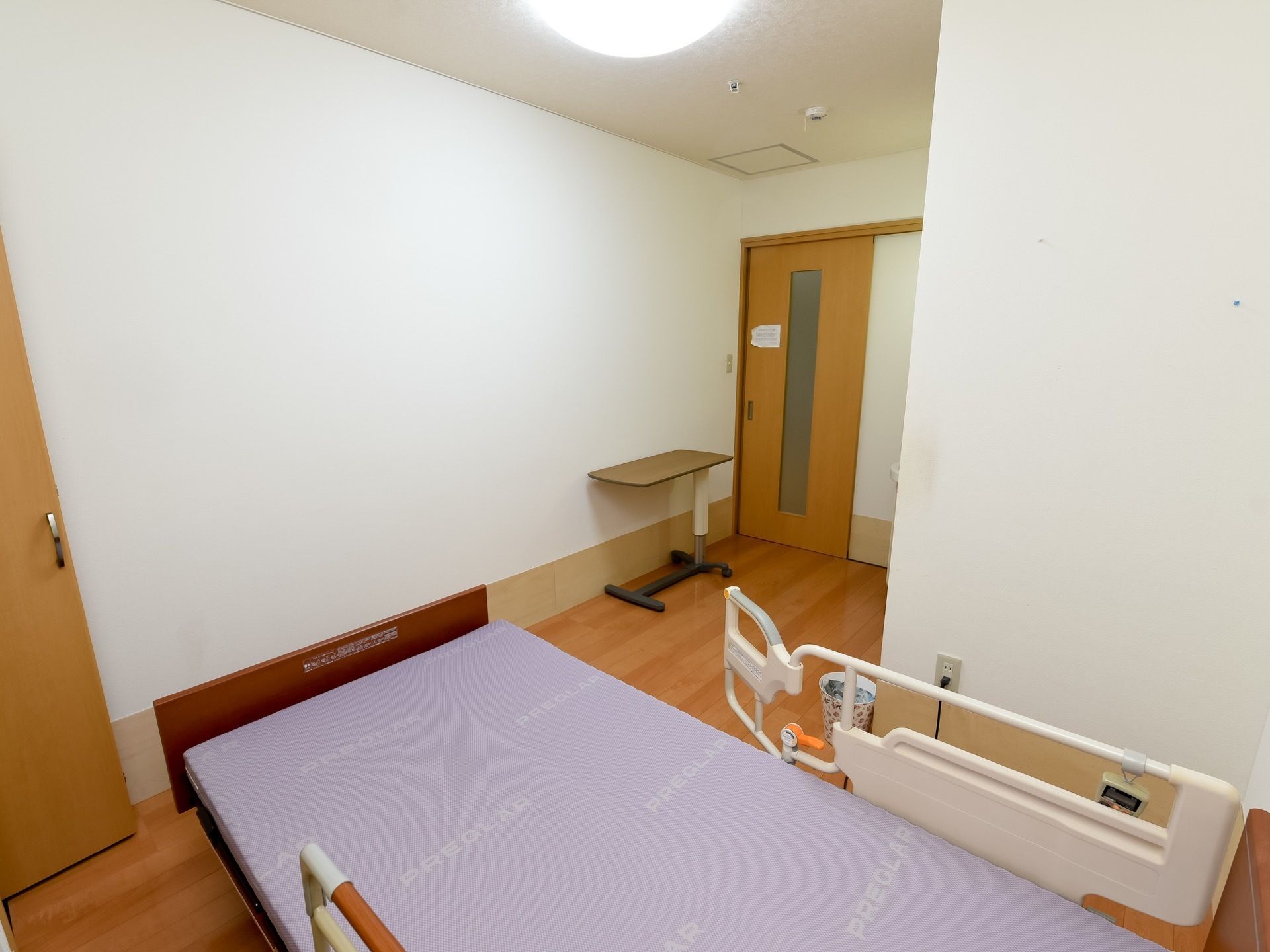 居室の入口前にテーブルが１台置かれている。落下防止の柵がついた介護用ベッドがあり、その横に収納の扉が開いている。