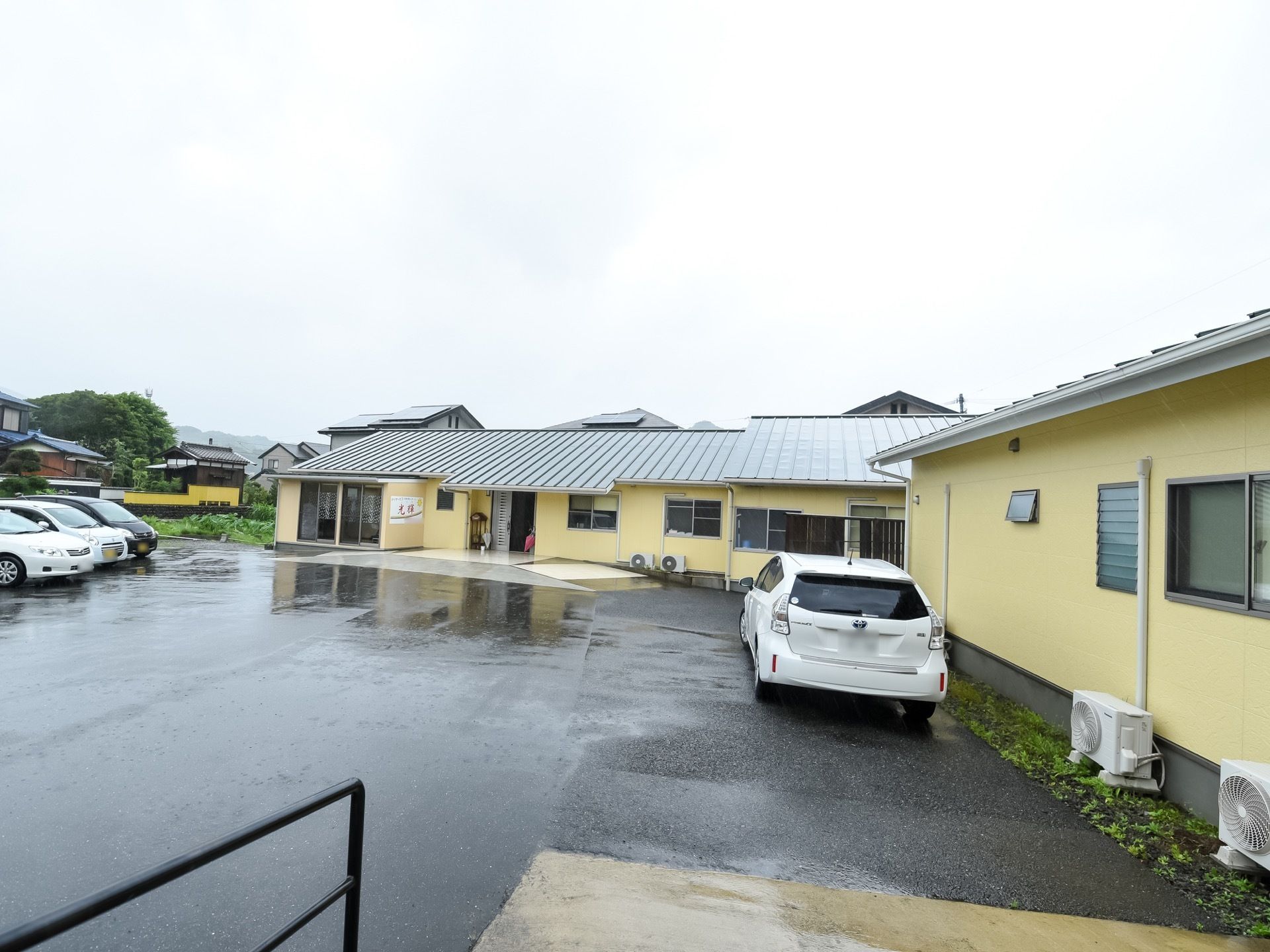 施設は平屋建てになっている。ベージュの壁に三角屋根がのっている。施設の前は駐車スペースになっている。