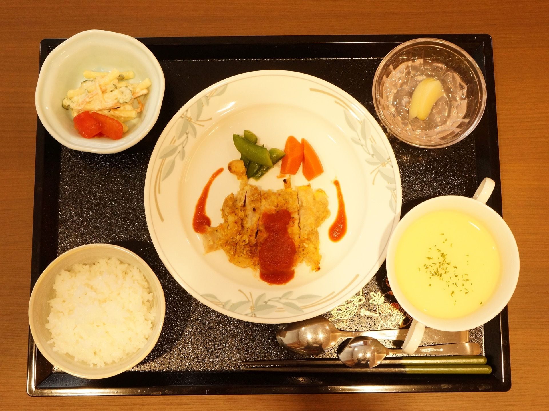 黒いお盆の上にはスープ用とデザート用のスプーンが2つとお箸、食事が配膳されている。ごはん、コーンスープ、カツ、サラダなどの献立である。