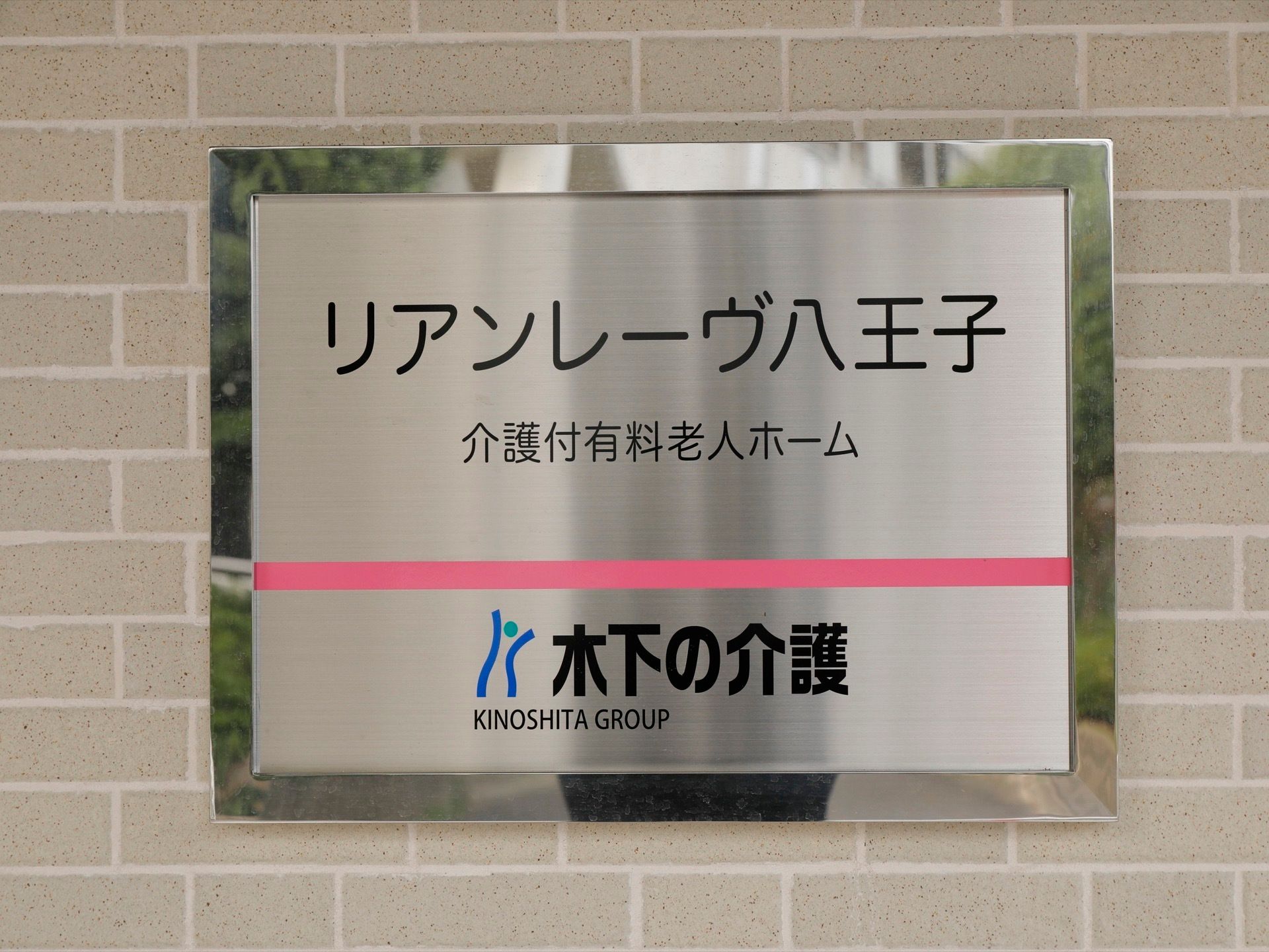 シルバーの枠組みのピンクのラインが入った看板となっている。読みやすい書体で施設名やグループ名が書かれている。