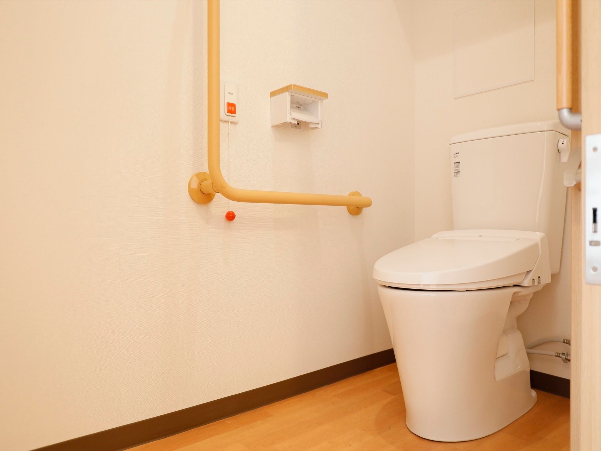 居室に備えつけられたトイレの壁にはＬ字型の手すりが備えつけられており、手すりの近くには呼び出しボタンも設置されている。