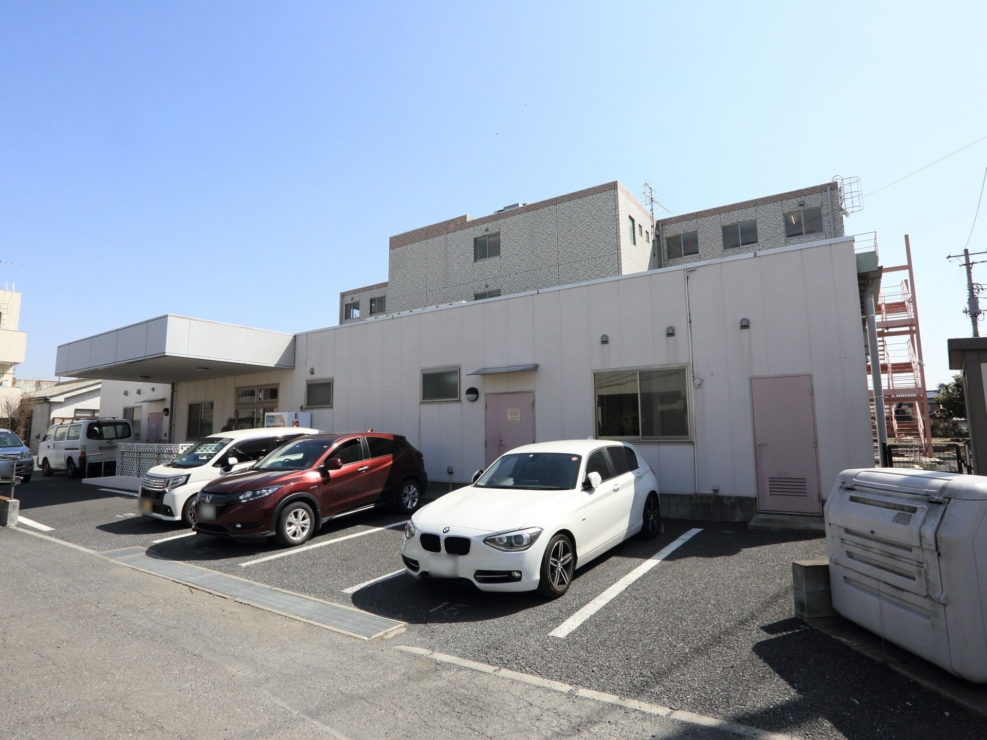 グループホームは４階建ての建物で、エントランスには自動販売機が置かれている。また敷地内には、駐車スペースが確保されている。