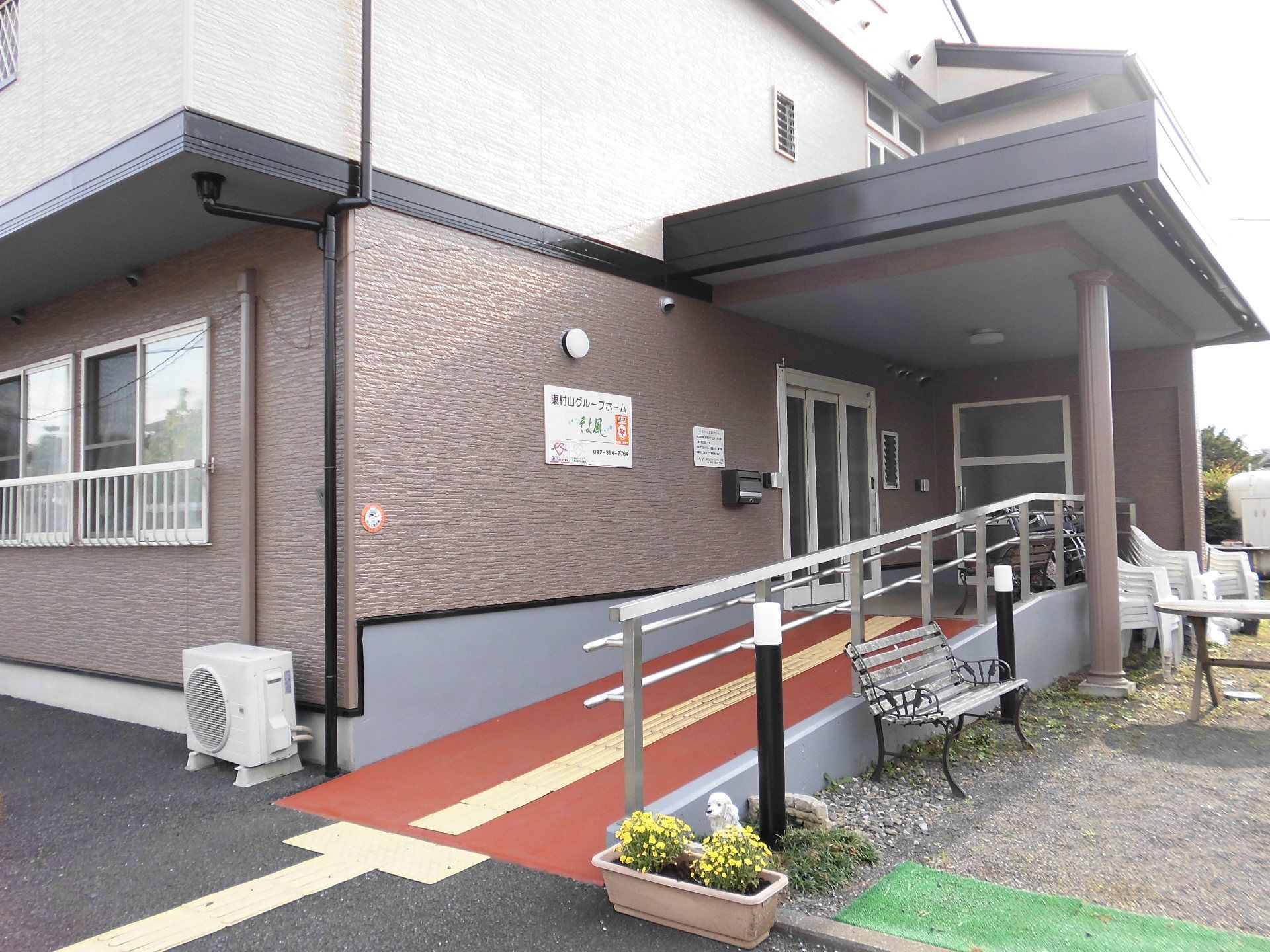 施設はブラウンが基調で落ち着いた雰囲気である。玄関までの通路は手すり付きのスロープになっており、車椅子をご利用の方も安全に移動できる。