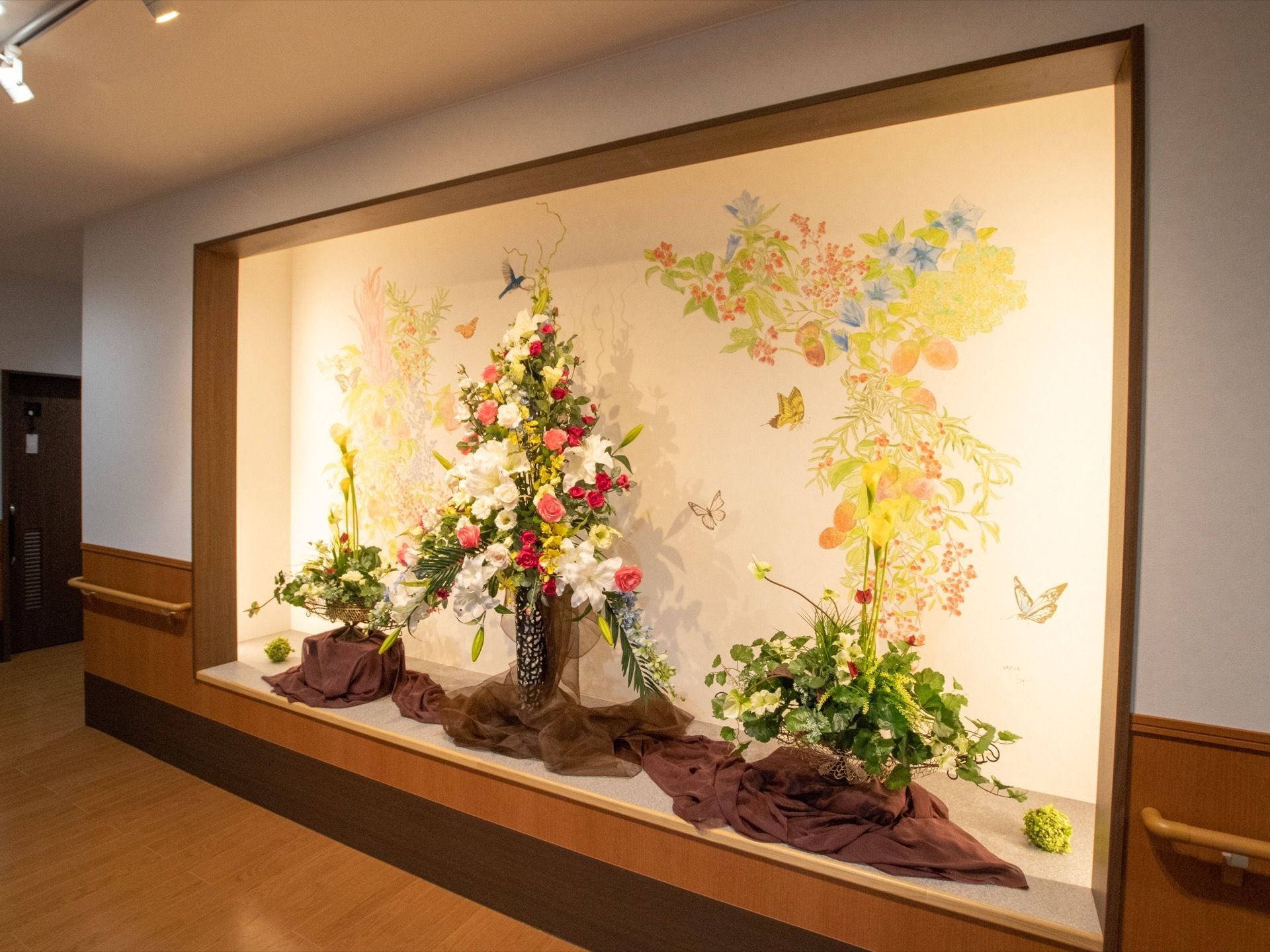 壁をくり抜いた大きなスペースに３つの生け花と壁画を組みあわせた作品が展示されている。奥には居室の入口が見える。