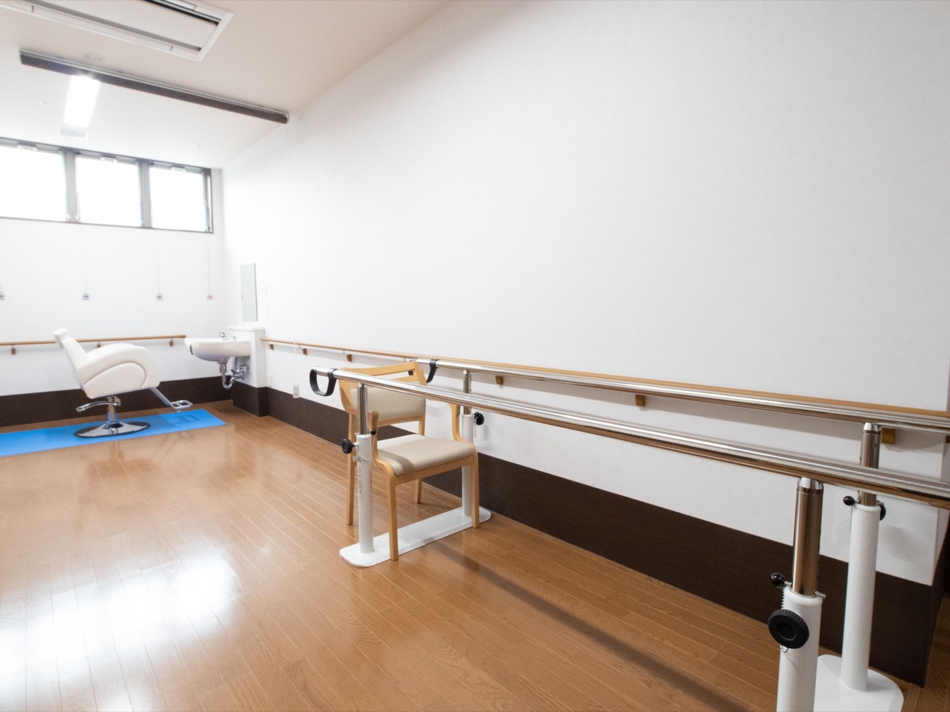 リハビリ用の平行棒があり、端に椅子が１脚置いてある。壁は白色で、茶色の手すりが設置されている。奥には理容スペースがある。