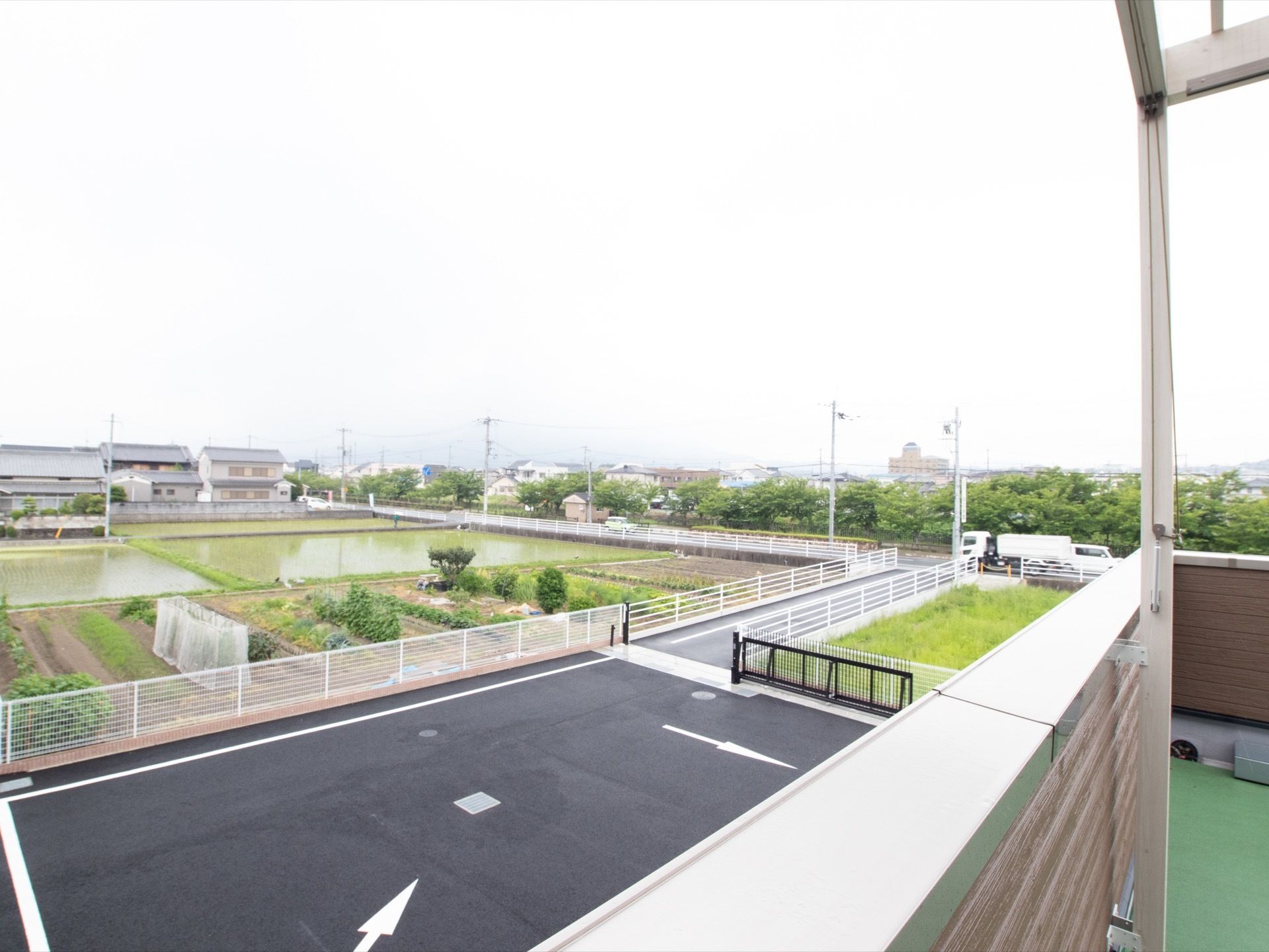 ベランダから見た周囲の景色の様子で、水田や畑が多くある。アスファルト舗装された部分には車誘導用の矢印が書かれている。