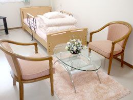 居室には窓が完備され、介護用ベッドが設置されている。置かれているテーブルセットは、明るい配色の椅子とガラステーブルである。