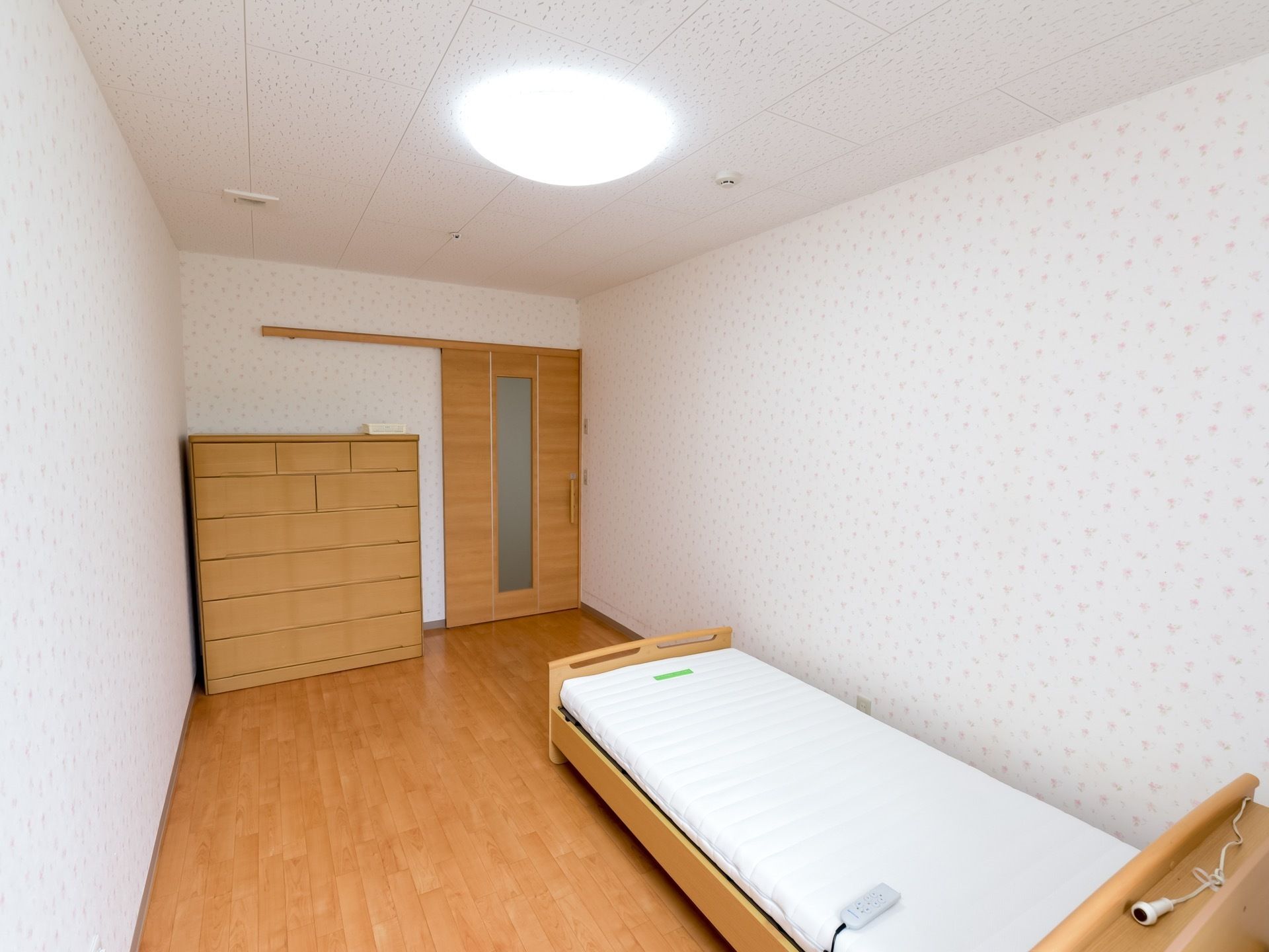フローリングの床に、介護用ベッドが置かれている。居室の扉は、木目調のスライド式ドアである。ベッドには、緊急時用のコールが置かれている。