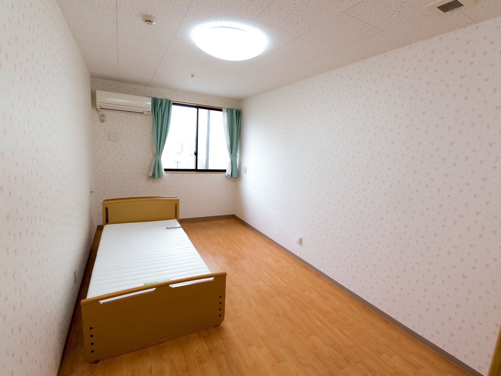 一人用の個室で壁側にベッドがおかれている。壁上部にはエアコンが設置されている。天井に火災報知器も備えつけられている。