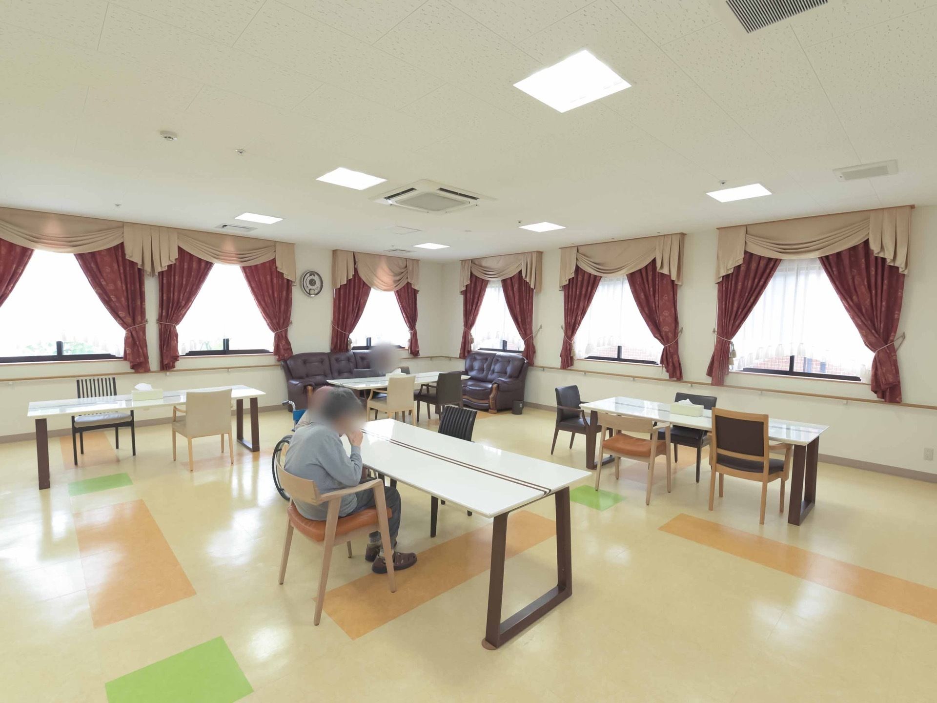 入居者様が椅子に腰をかけテーブルでお茶を飲まれて過ごされている。複数の窓が設置さておりお洒落なカーテンが掛けられている。