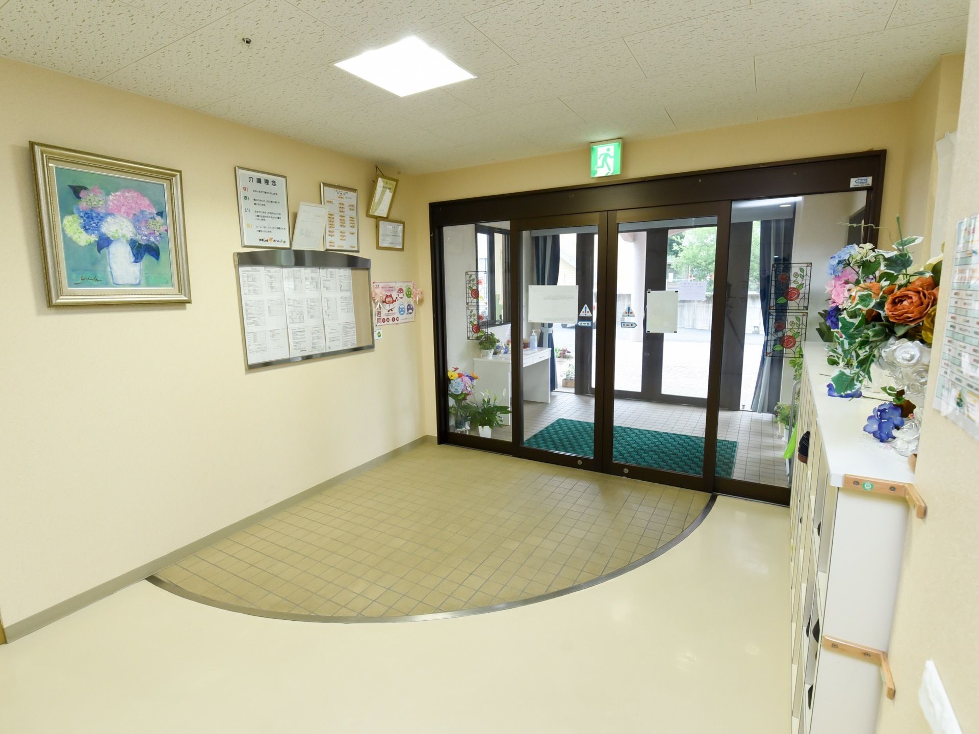 ガラス張りの自動ドアから外光が差し込む明るいエントランスホールである。右端に靴箱があり、その上に花が飾ってある。
