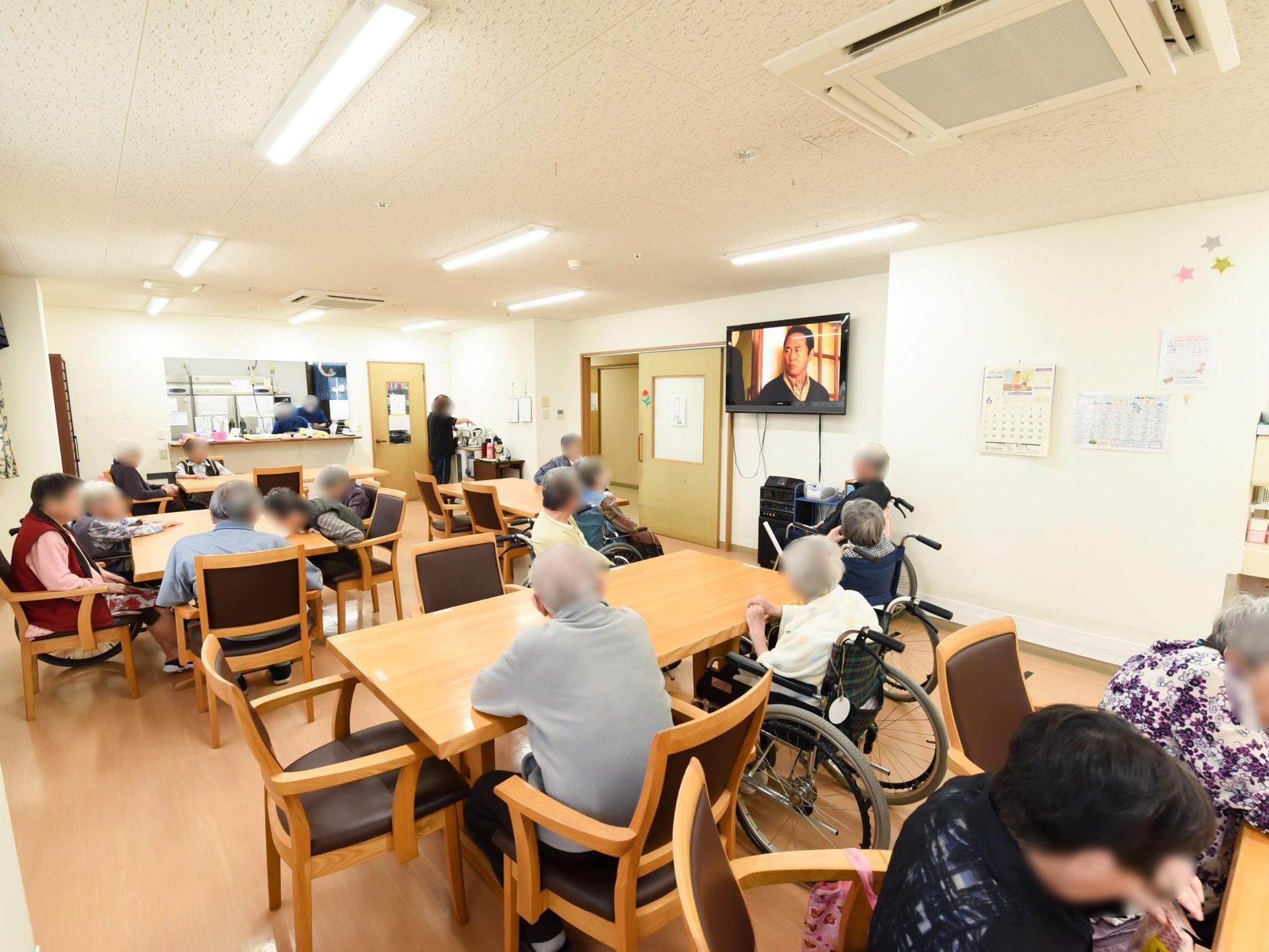 テーブルの周りに数多くの入居者様が座っていて、テレビを見ながらくつろいでいる。右側の壁には、カレンダーが掛けてある。