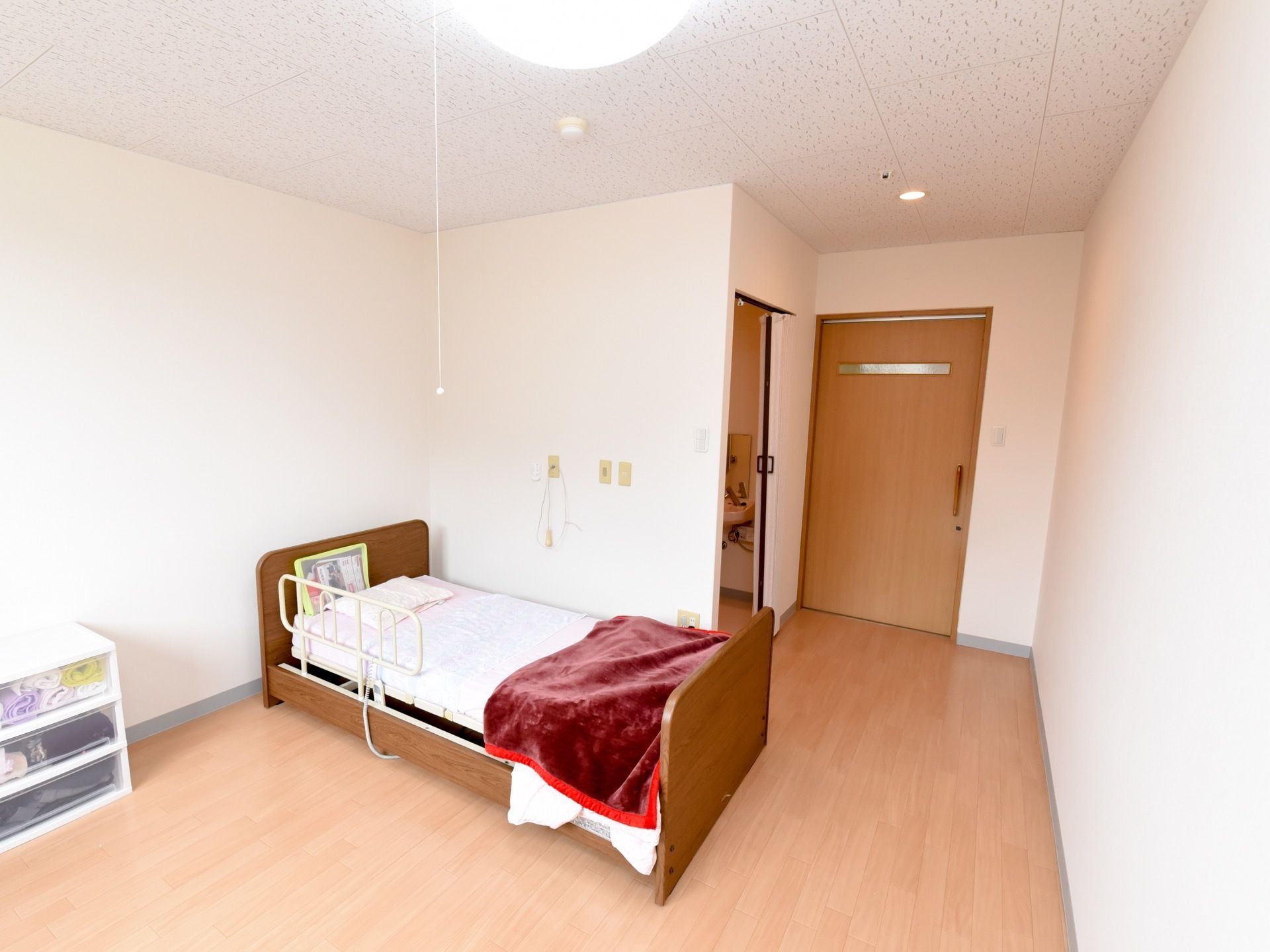 白い壁とライトブラウンの床が、すっきりとしたシンプルなデザインの居室である。壁際には、収納ケースや電動ベッドが置いてある。