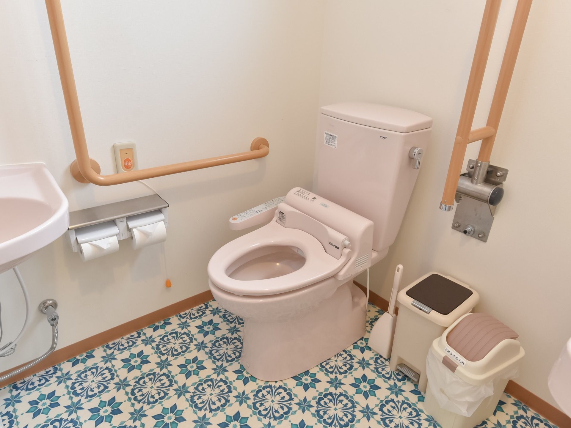 ピンクの温水洗浄便座が設置され、壁には木目調の手すりや呼び出しボタン、トイレットペーパーが備えられている。