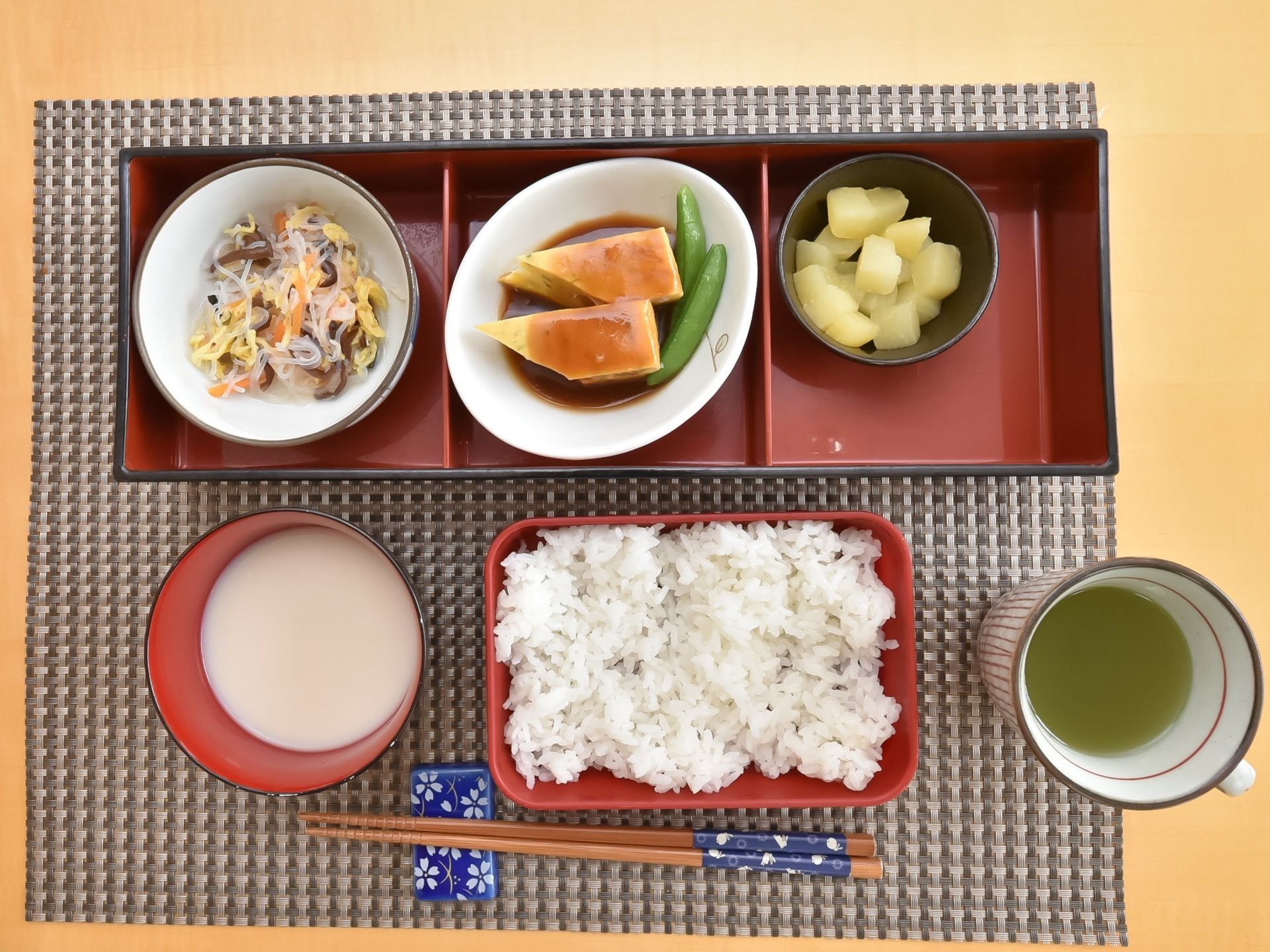 花柄の青い箸置きにお箸がおいてある。赤くて四角い器にご飯が入れられており、長方形の仕切りが付いた器には煮物などが並べられている。