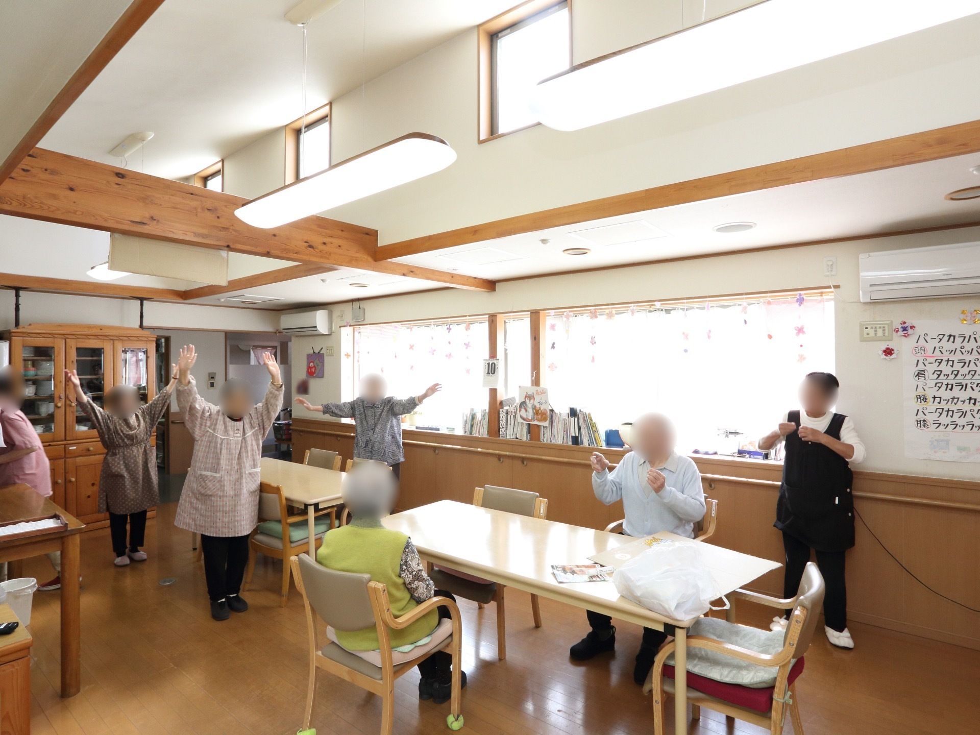 スタッフと一緒に入居者様が手を挙げ体を動かしている。食堂の入口付近には、木製の茶箪笥が置かれている。