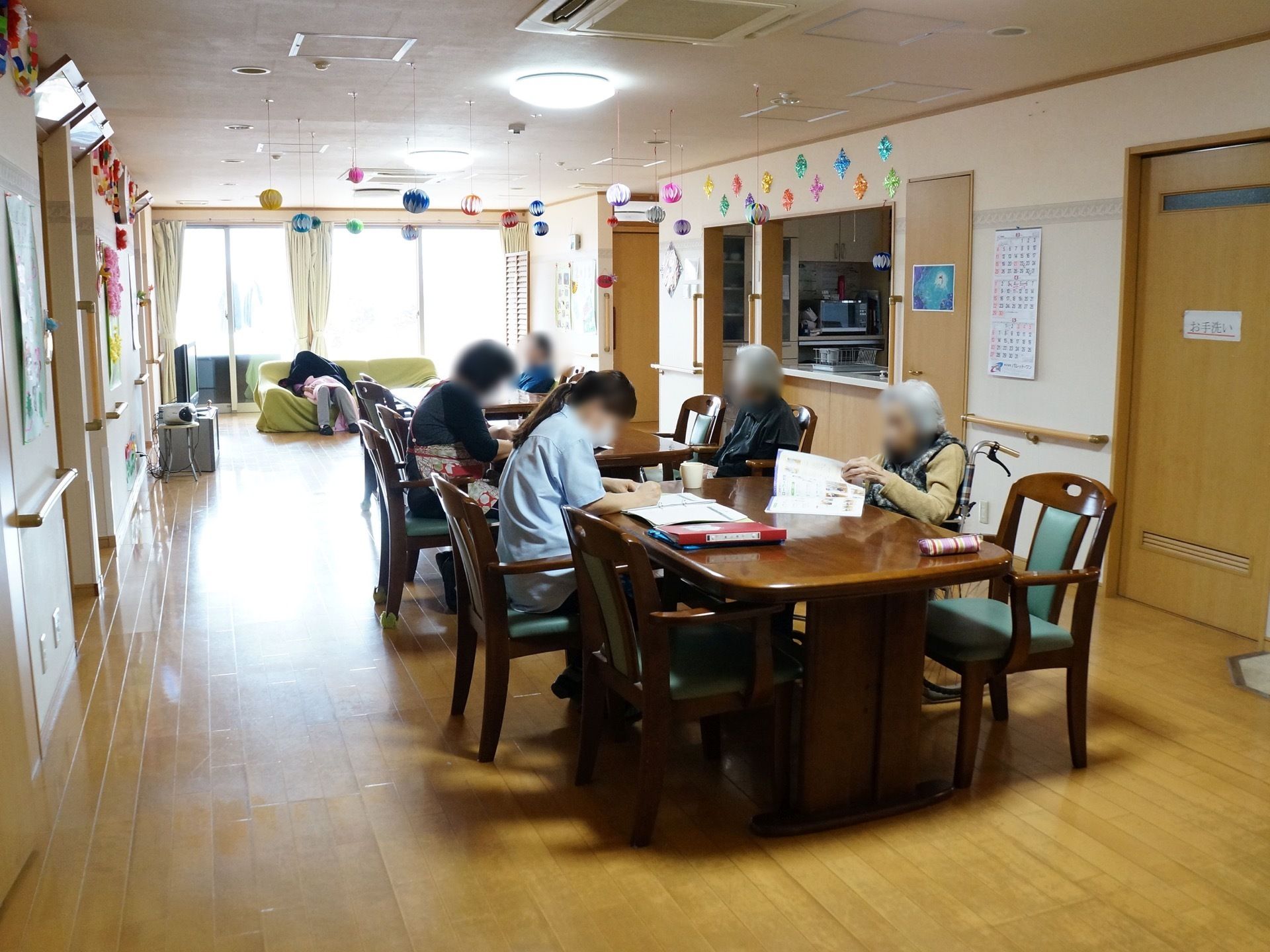 大きな窓が設置された共有スペースである。日差しが入り明るい。配備されたテーブルでは、スタッフと入居者様が同じ場所で、時間を共有している。