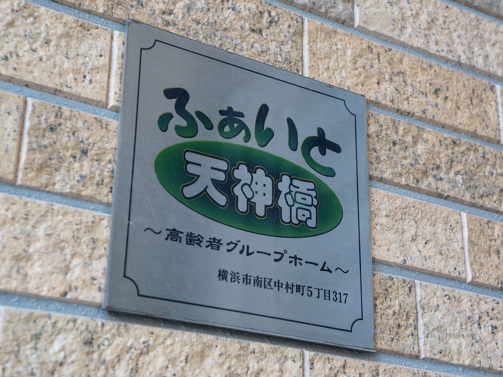 施設の壁には、施設名称、施設種別、住所が、黒と緑色のデザイン文字で描かれた、シルバーのプレートが貼られている。