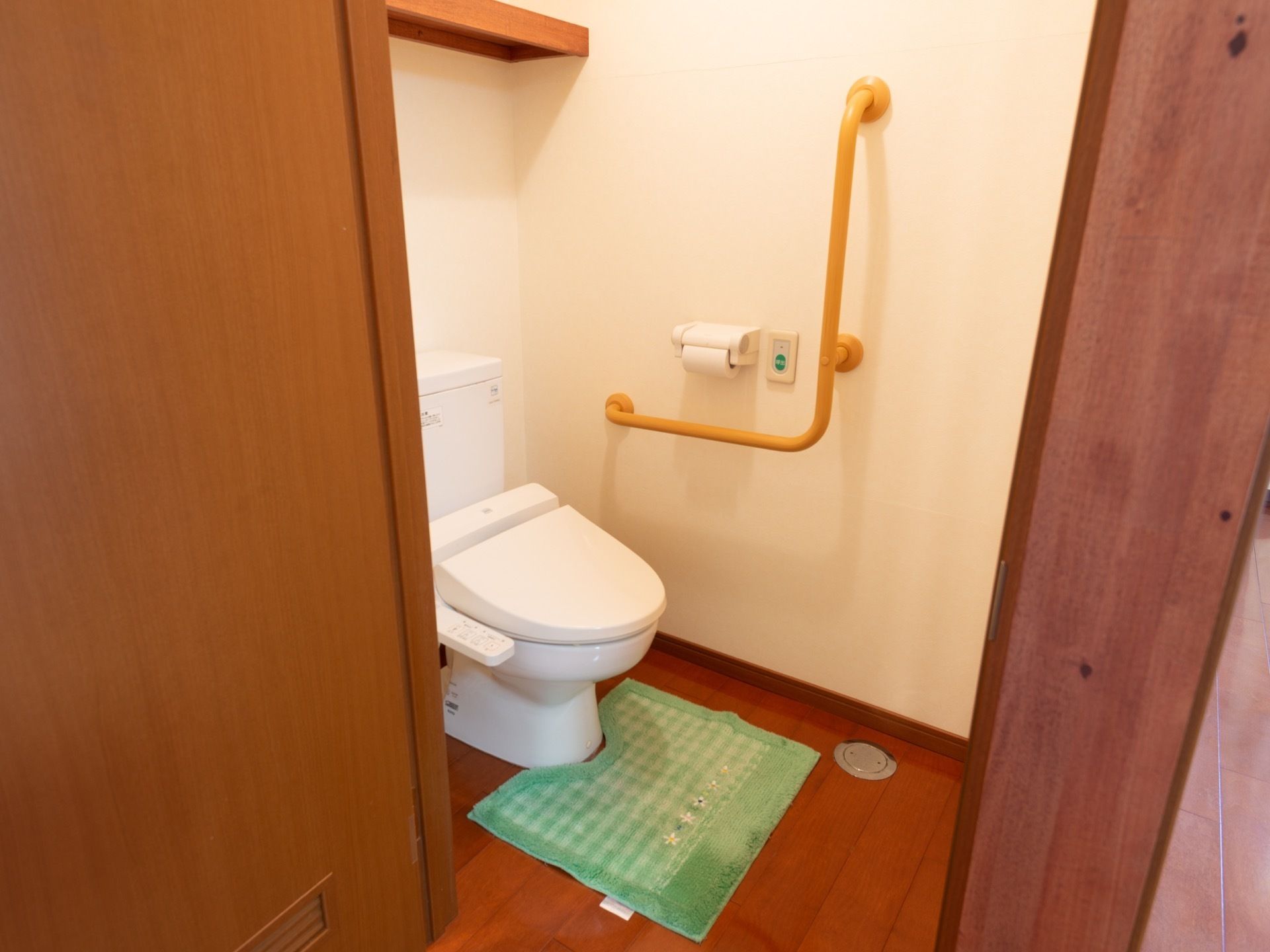 居室のトイレは、開き戸の中に設置されている。緑色のマットが敷かれている。貯水タンクの上部には棚板が取りつけられている。