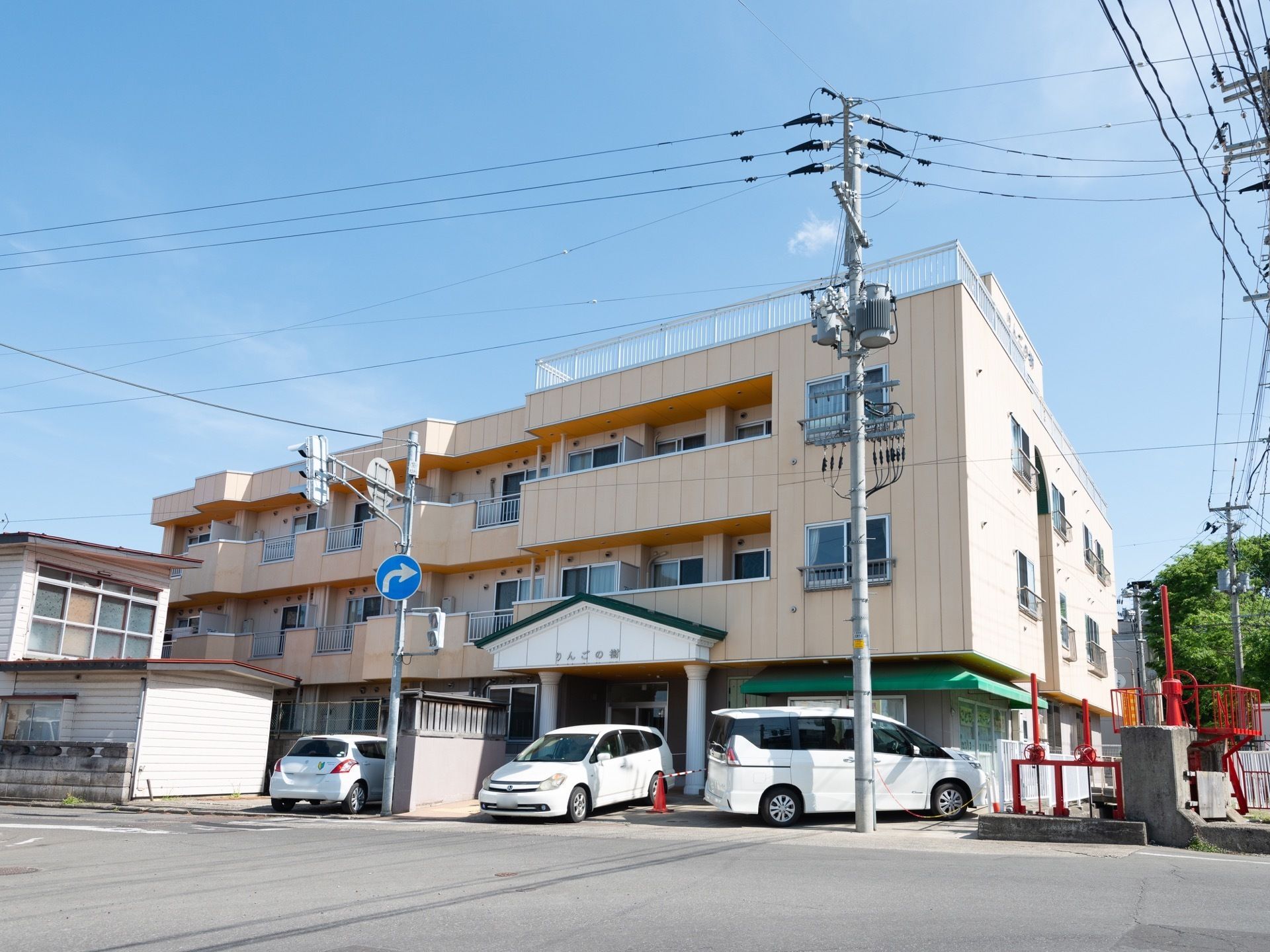 ３階建てのマンションタイプの建物がある。入口付近には乗用車が２台停まっている。前の車道は道幅が広くなっている。