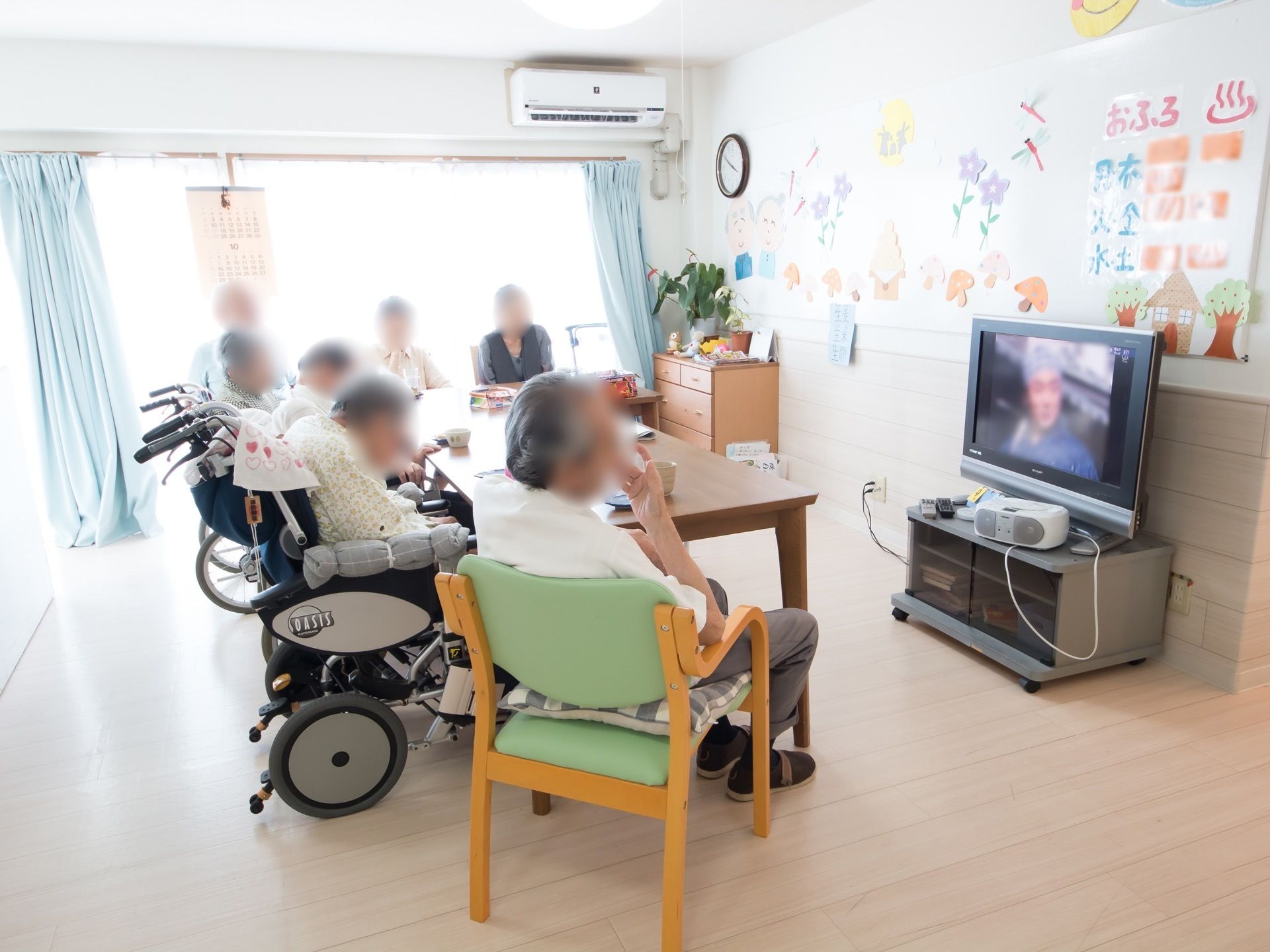 テレビの前には大きなテーブルがあり、入居者様たちが取り囲むように座っている。壁には季節感のあるペーパークラフトなどが飾られている。
