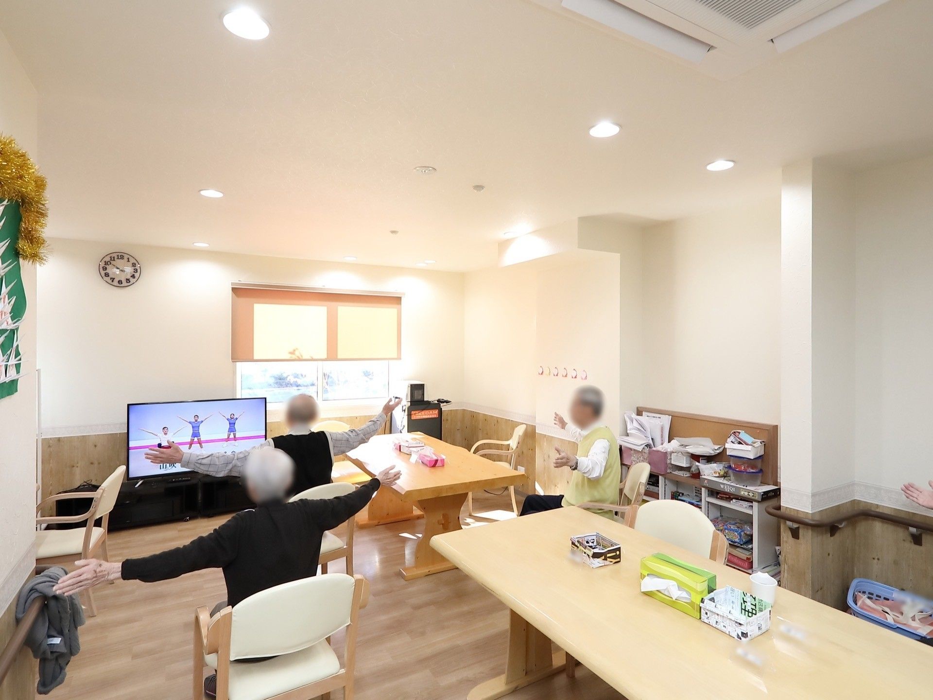 木のぬくもりあふれるリビングスペースで、ナチュラルブラウンの家具があたたかみを感じさせる。入居者様がテレビを観ながら、体操をしている。