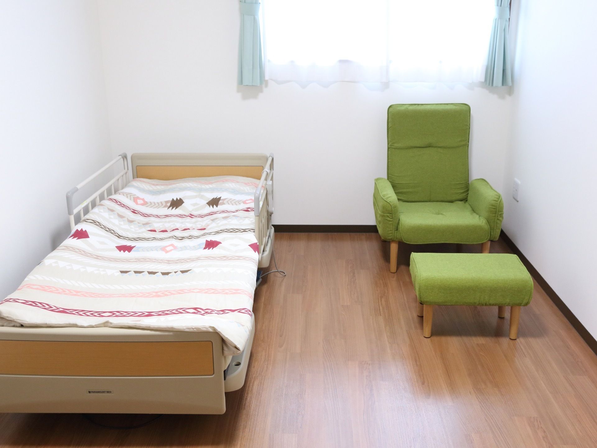 陽当たりの良い窓に面した居室。床はフローリングになっている。ベッド、オットマン付きの椅子が置かれている。