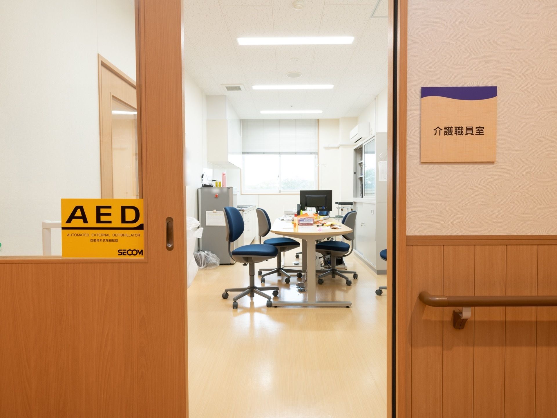 スタッフの事務所入口横に看板が貼られている。入口扉の窓にAEDの表示がされている。室内にテーブルと椅子が並んでいる。