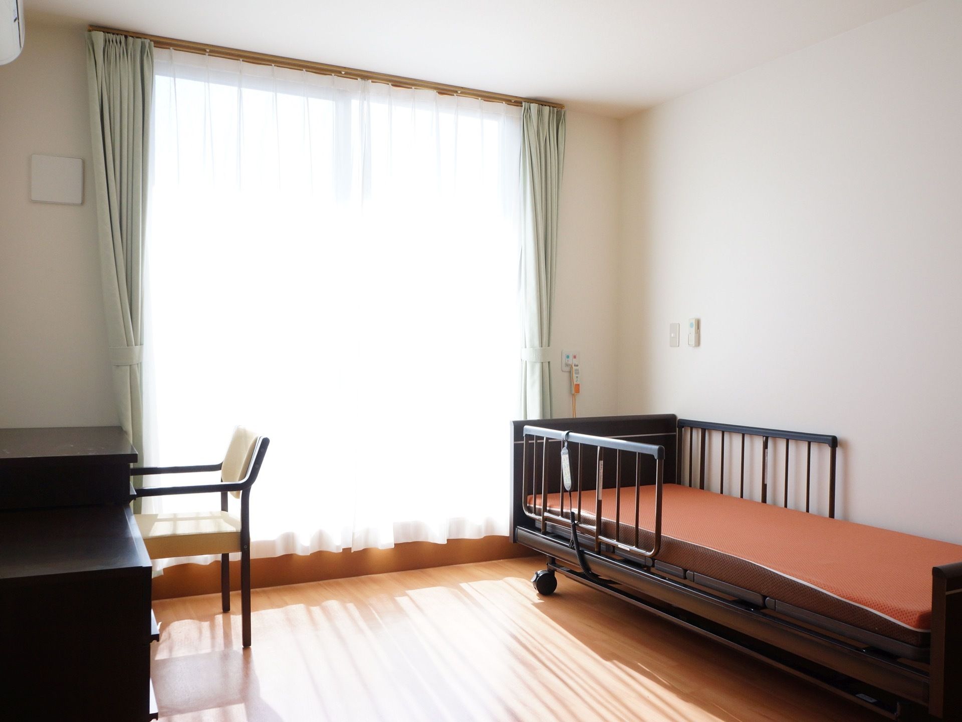 電動式介護ベッドが、居室の右側に設置されている。すぐ側にナースコールのボタンがある。左側には、デスクと椅子がある。