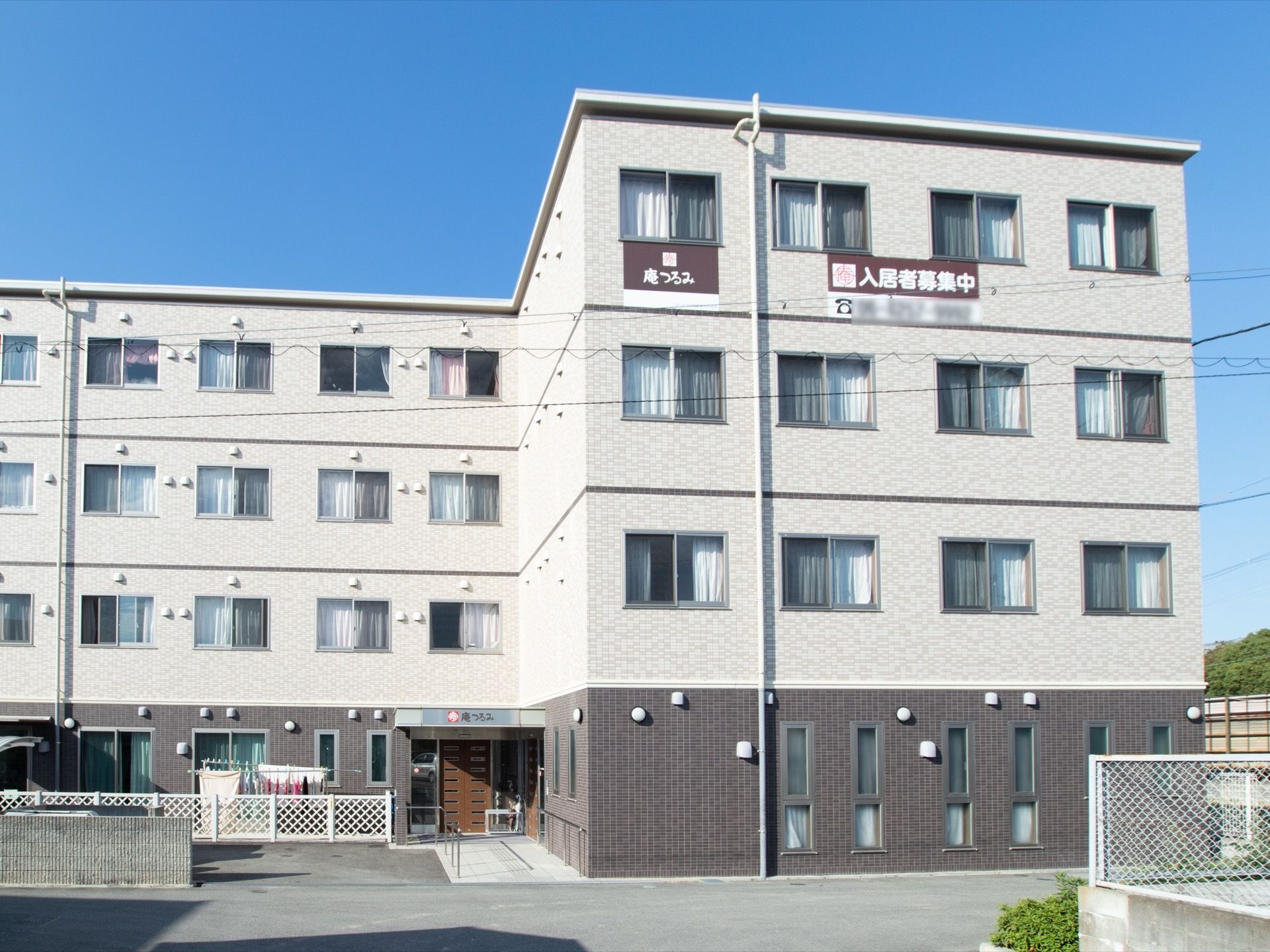 施設は４階建てで、１階がグレー、２階より上がホワイトの外観である。白いフェンス内には洗濯物が干されている。