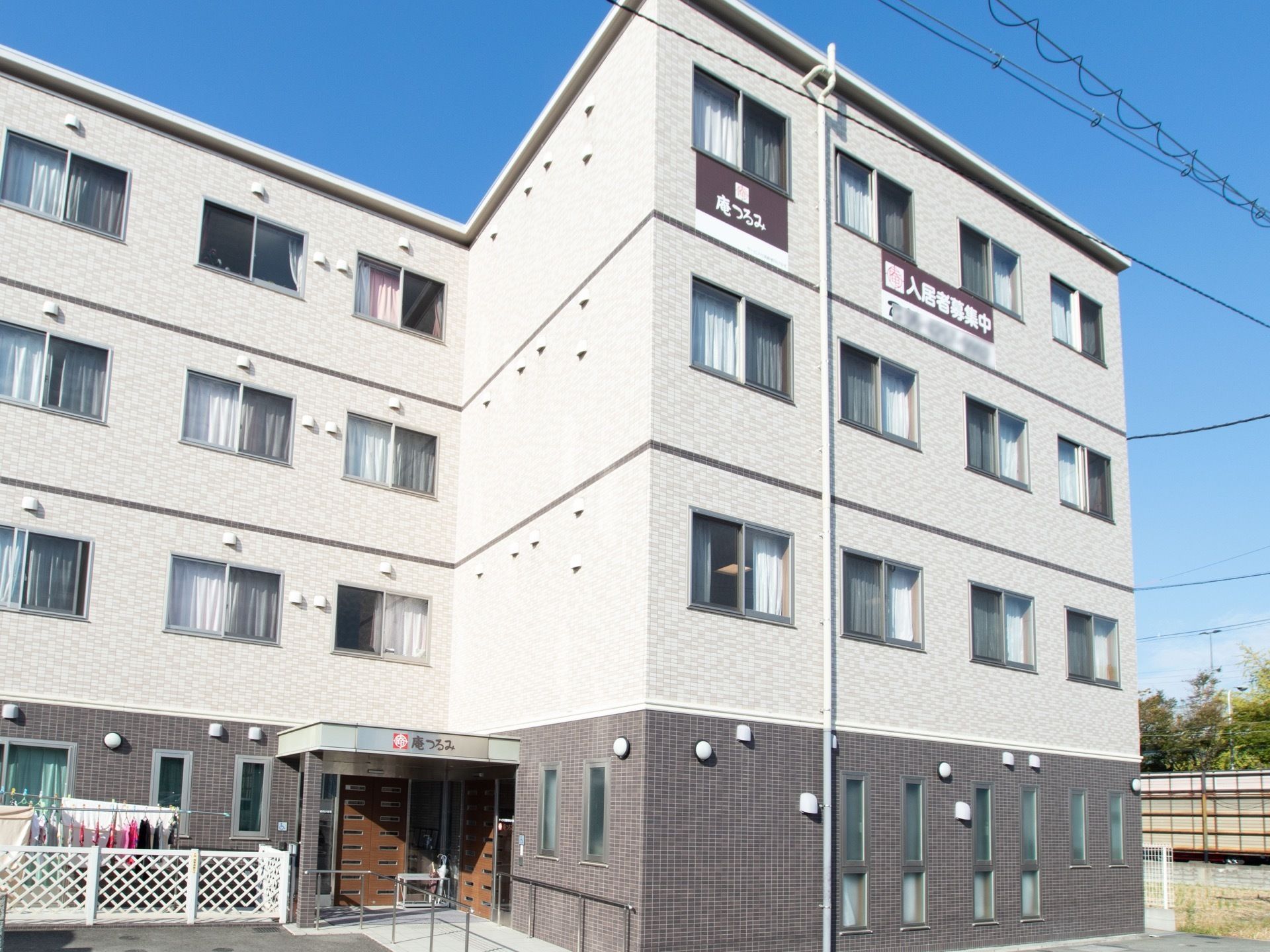 施設は４階建てである。玄関前にはスロープがあり、両側に手すりが取りつけられている。外壁には入居者様募集の看板が掲げられている。