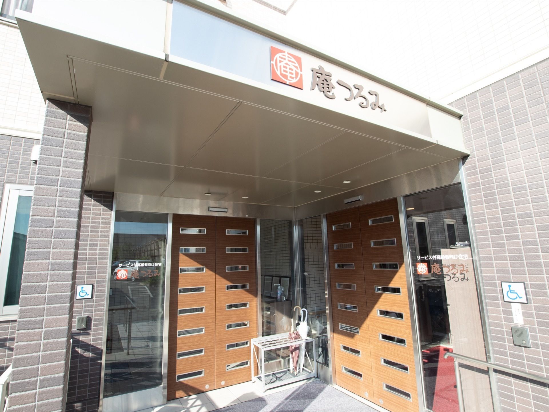 玄関には木製の扉が２つ設置されている。前には傘が入った傘立てがある。大きな屋根があり、施設名が書かれている。