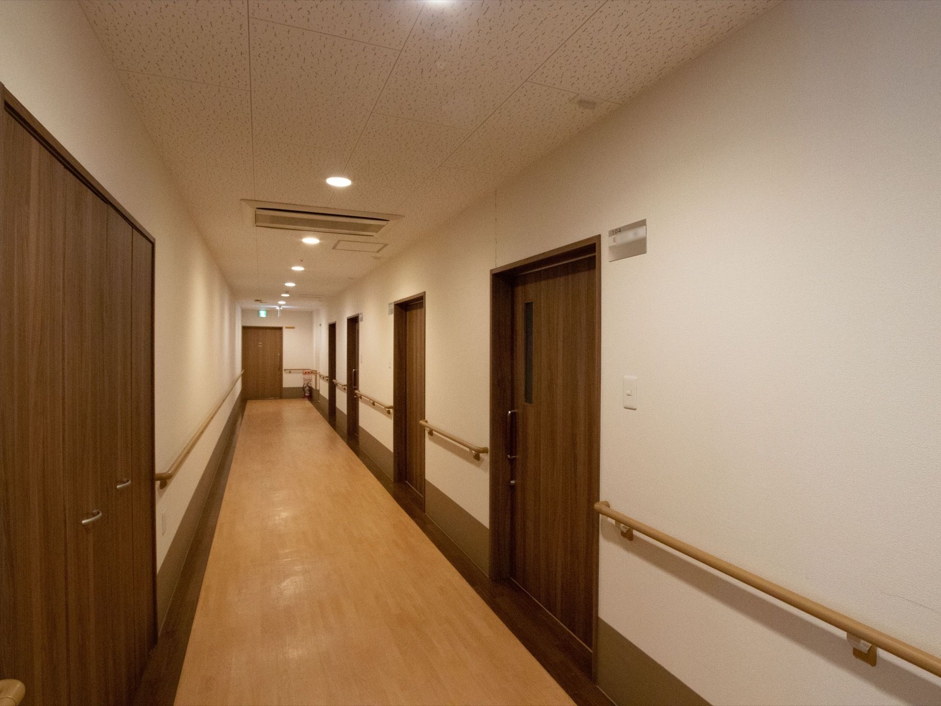 まっすぐに続く通路がある。通路両側には部屋の扉と手すりが取りつけられている。天井には避難経路誘導灯もある。