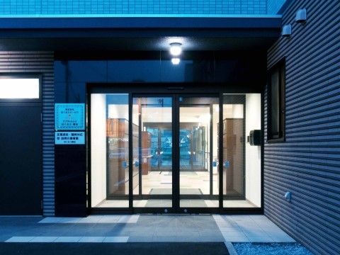 中央にガラス張りの自動ドアがあり、左の壁に施設名の書かれたプレートが貼ってある。玄関の左には勝手口があり、上の小窓から明かりが漏れている。