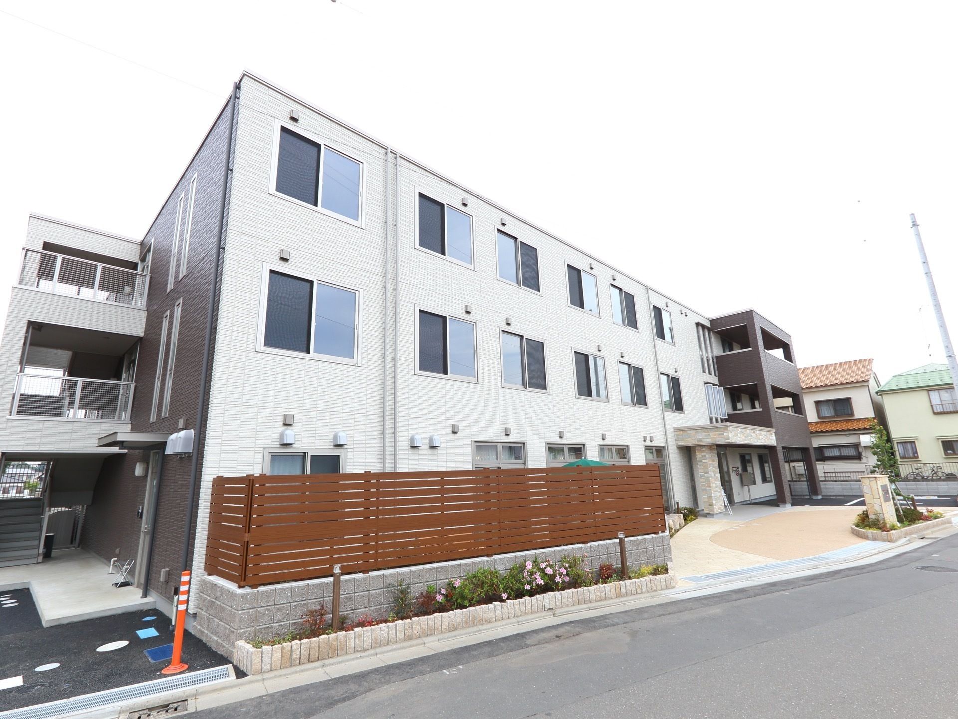 3階建てのモダンな建物である。植え込みには色鮮やかな花が咲いていて美しい。目隠しのための茶色い塀が建てられている。
