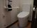 Ｌ型手すりや呼び出しボタンが完備されたトイレである。足元には、トイレマットが敷かれている。扉は引き戸になっている。