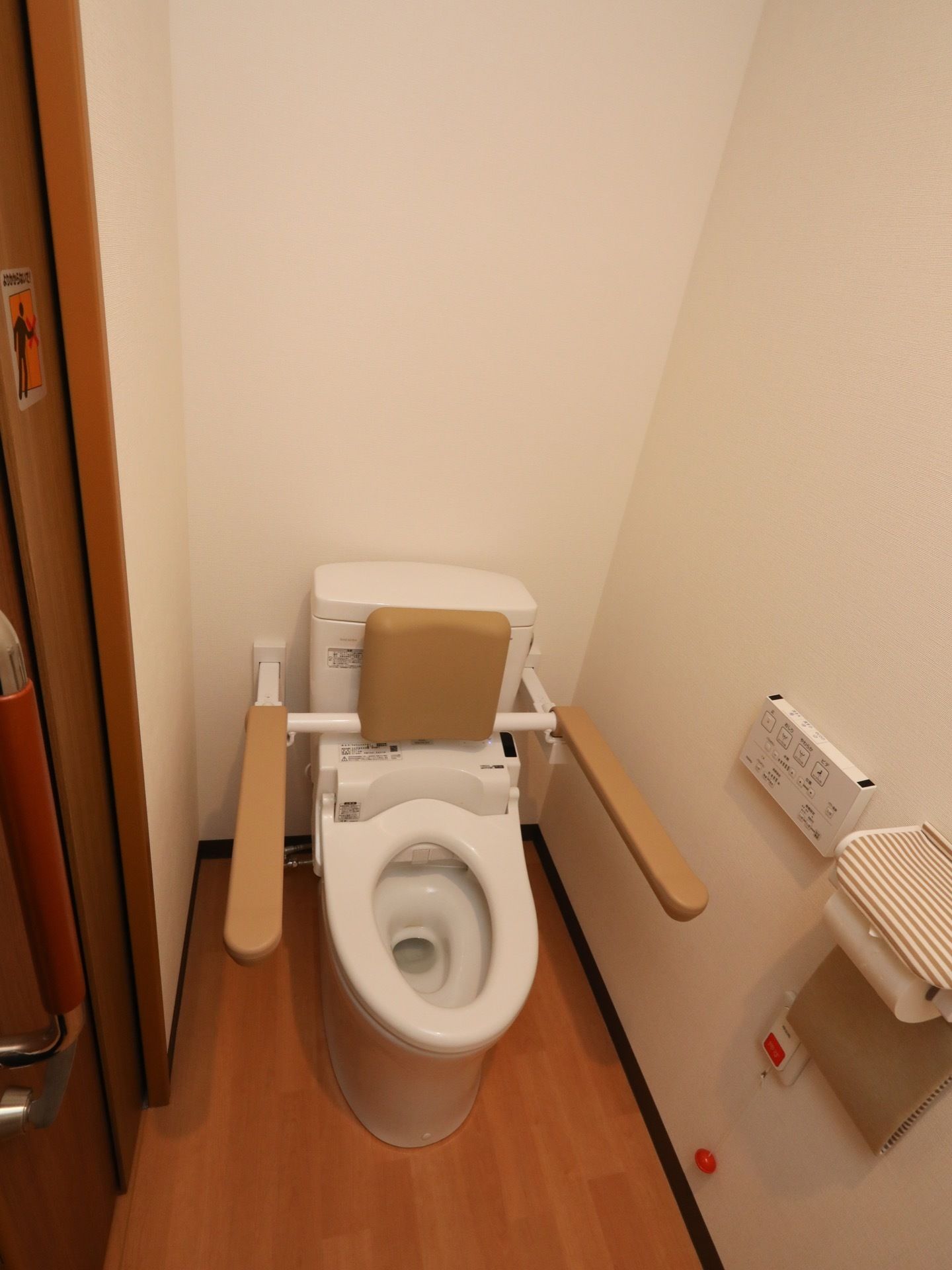 トイレの便座の周囲に、背もたれやひじかけが設置されている。阿部の下の方に、オレンジ色の玉がぶら下がった呼び出しボタンがある。