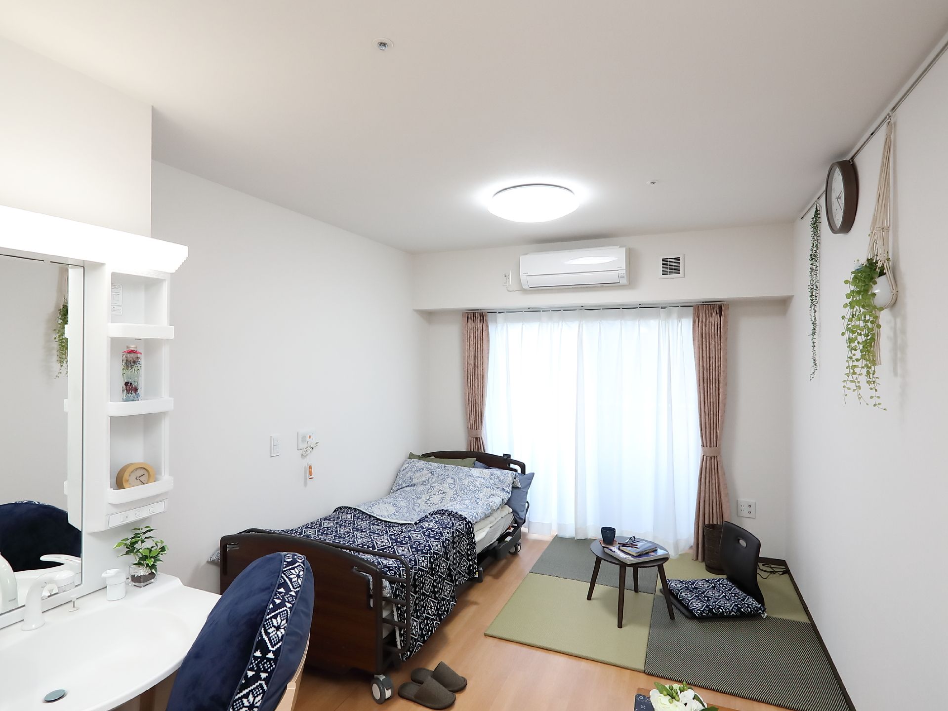 エアコンや洗面所が付いた居室はフローリングで落ち着いた雰囲気。机セットや観葉植物、ベッドが置かれている。