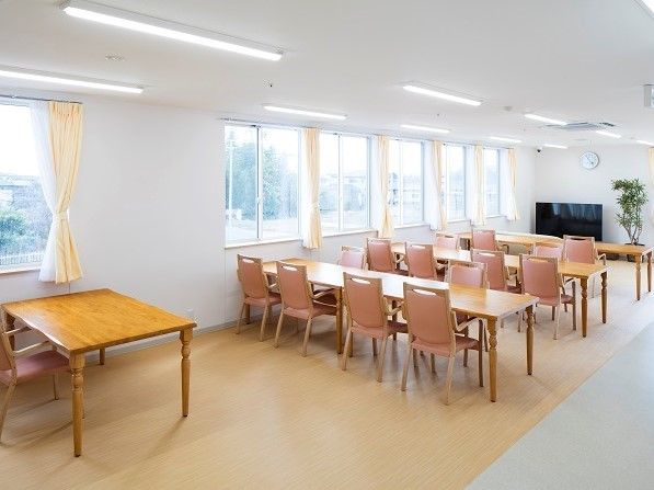 窓と蛍光灯が複数設置された明るい空間。長テーブルと椅子が複数並べられており、前方にはテレビと観葉植物が置かれている。