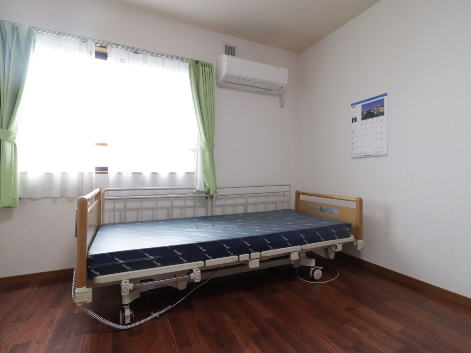 電動介護ベッドが備わっている居室である。レースのカーテンを通った柔らかい光が差し込んでいる。フローリングなので、家庭的な温もりを感じる。