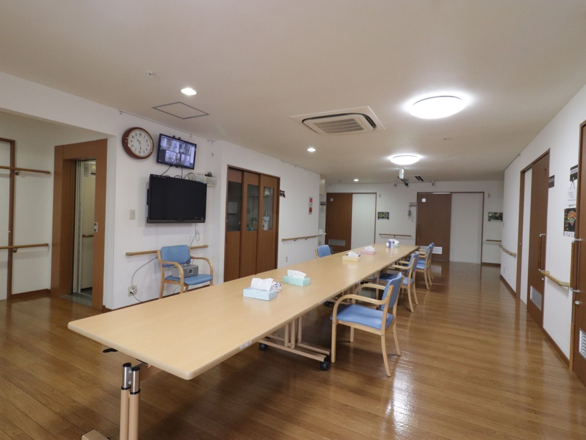 壁掛けテレビの前に長テーブルが並べられており、クッション付きの椅子が置かれた部屋