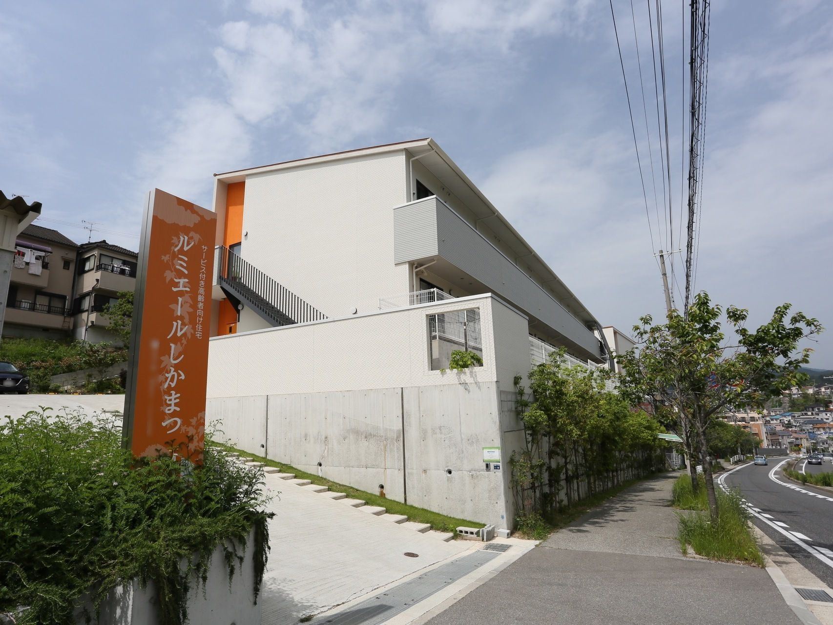 施設は丘の上にあり、坂道の下の方を見ると市街地が広がっているのが見える。歩道に接している部分は植え込みになっている。