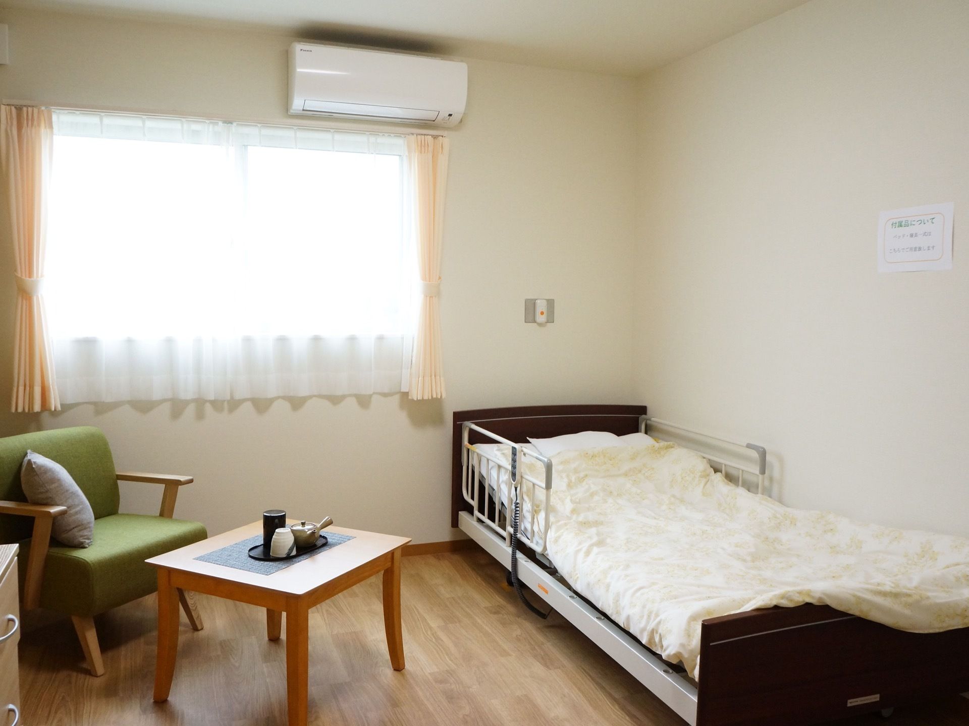 窓がある明るい居室には、電動介護ベッド、一人掛けソファとテーブルが置かれている。壁にはエアコンが設置されている。