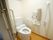 手前にある白い引き戸が左に引かれ、フローリングの部屋にトイレがある。奥の壁には手すりのほか、温水洗浄便座の操作パネルなどがある。